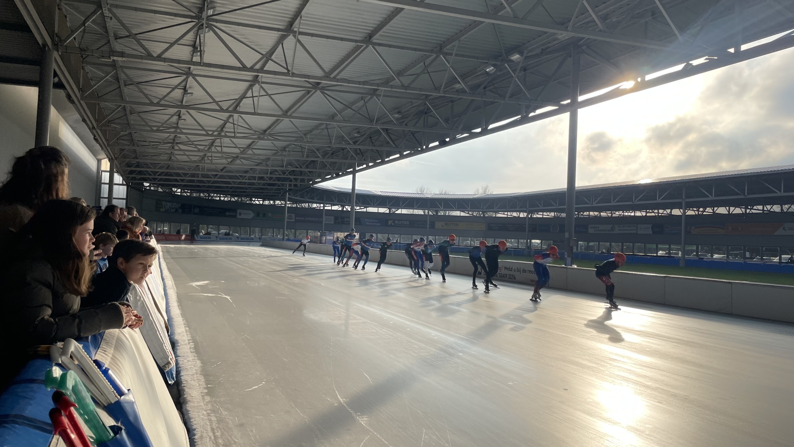Schaatsers racen op een overdekte ijsbaan, omringd door toeschouwers langs de zijkant.