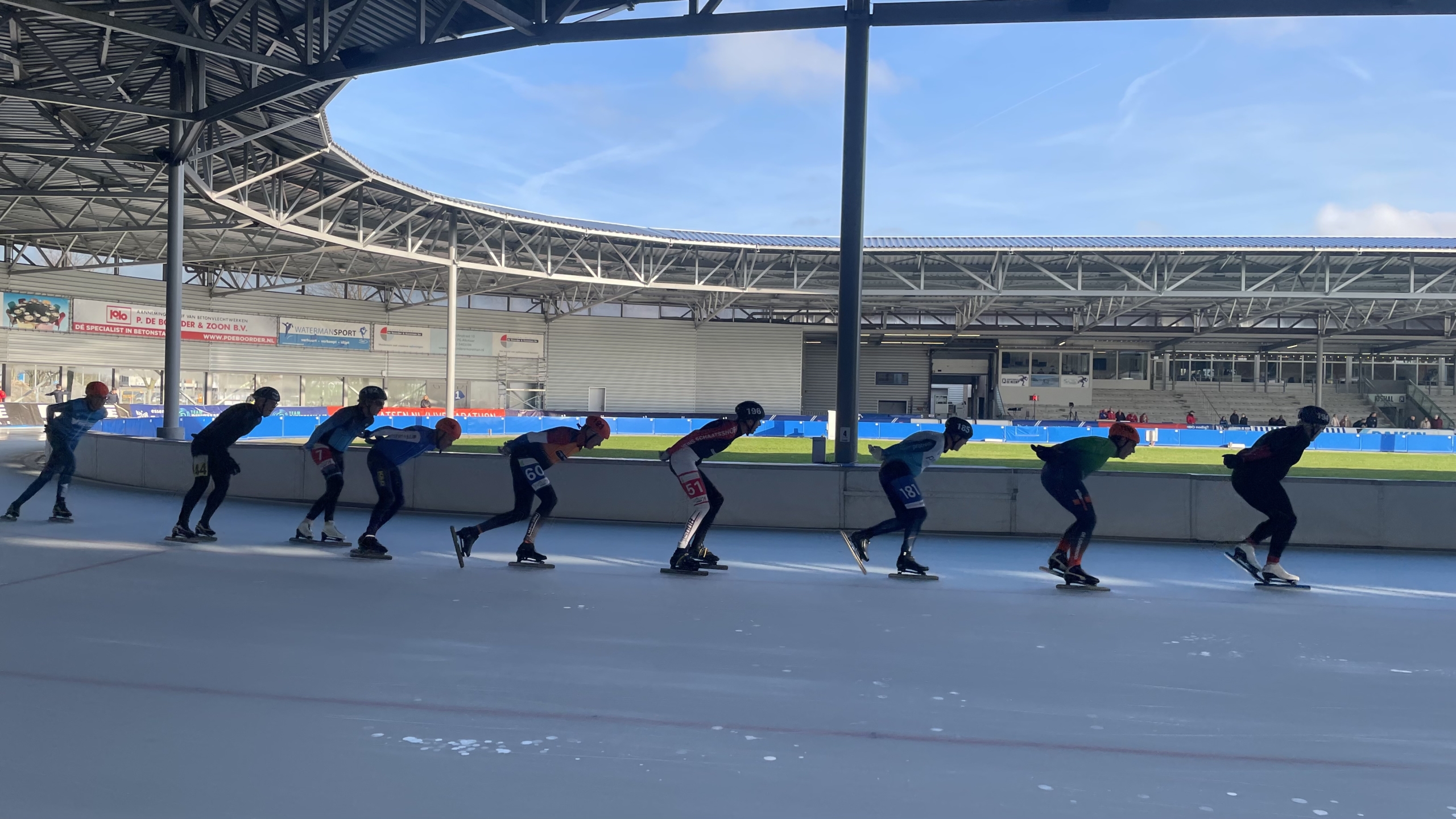 Schaatsers in competitieformaat op een ijshockeybaan, verlicht door een zonnige dag.
