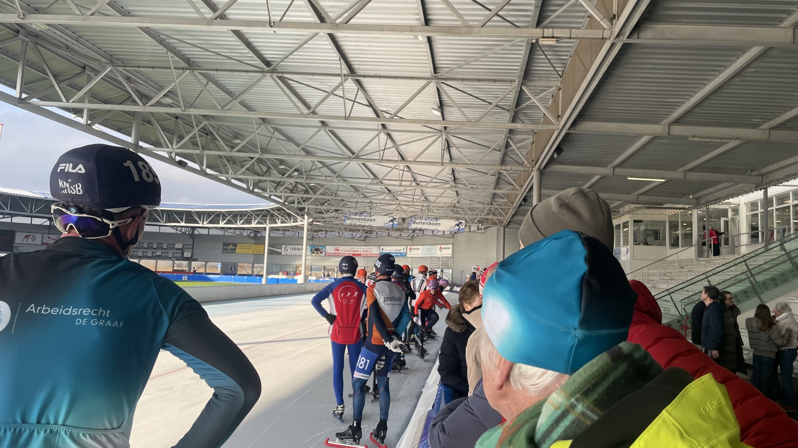 Schaatsers staan in een rij op een overdekte ijsbaan, omringd door toeschouwers.