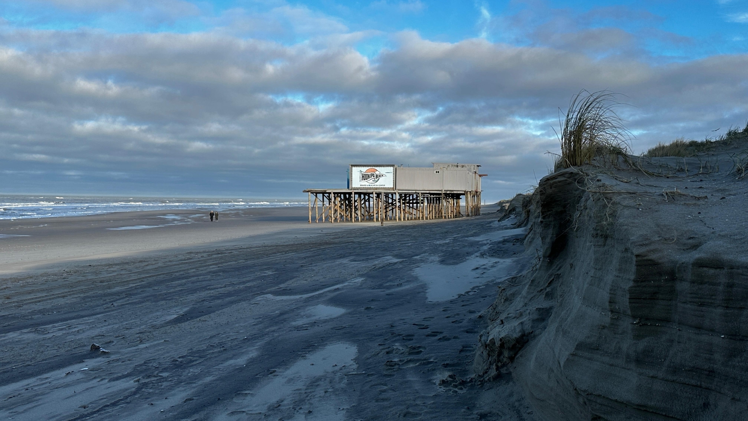 Strand met houten gebouw op palen, wolkenlucht en duinen aan de rechterkant.