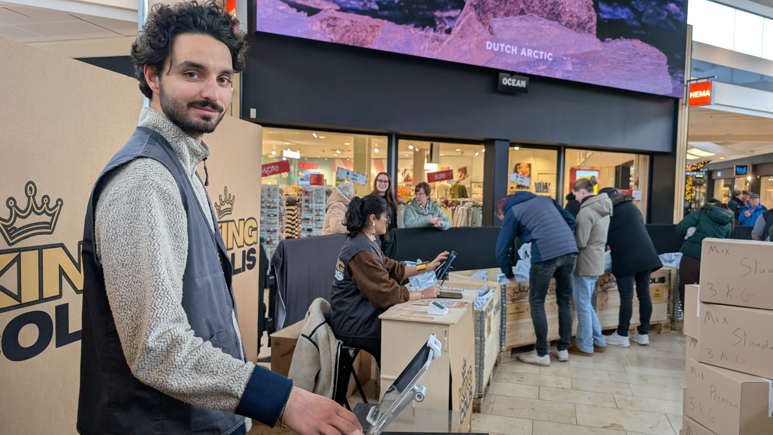 Man en vrouw bij een verkoopstand in een winkelcentrum, met mensen die dozen bekijken.