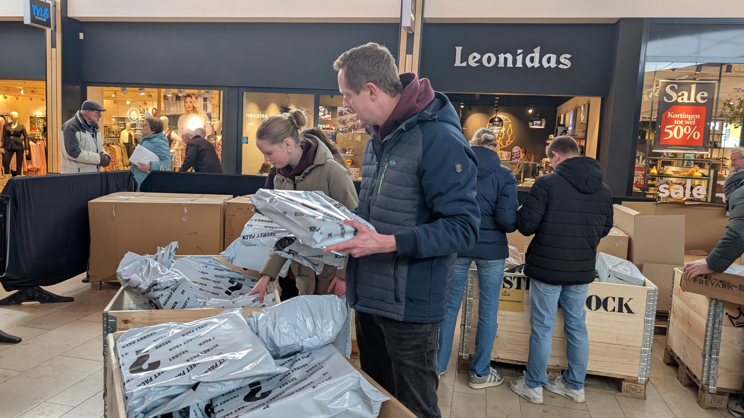Mensen bladeren door dozen met plastic verpakte producten in een winkel met een Leonidas winkel op de achtergrond.