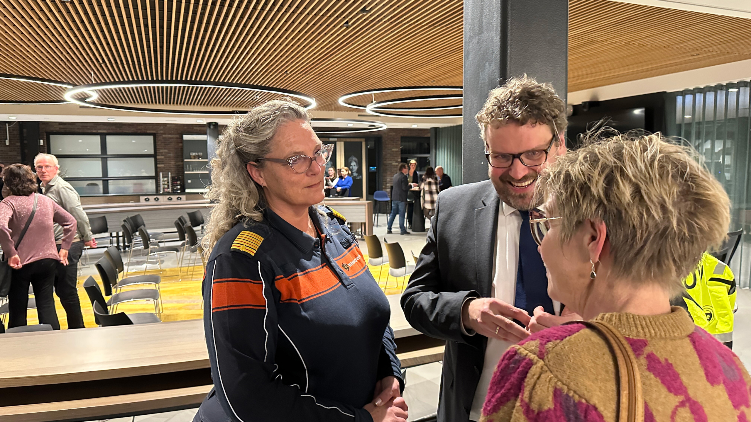 Drie mensen in gesprek in een moderne hal met houten plafond en stoelen; één draagt een uniform met oranje en navy strepen.