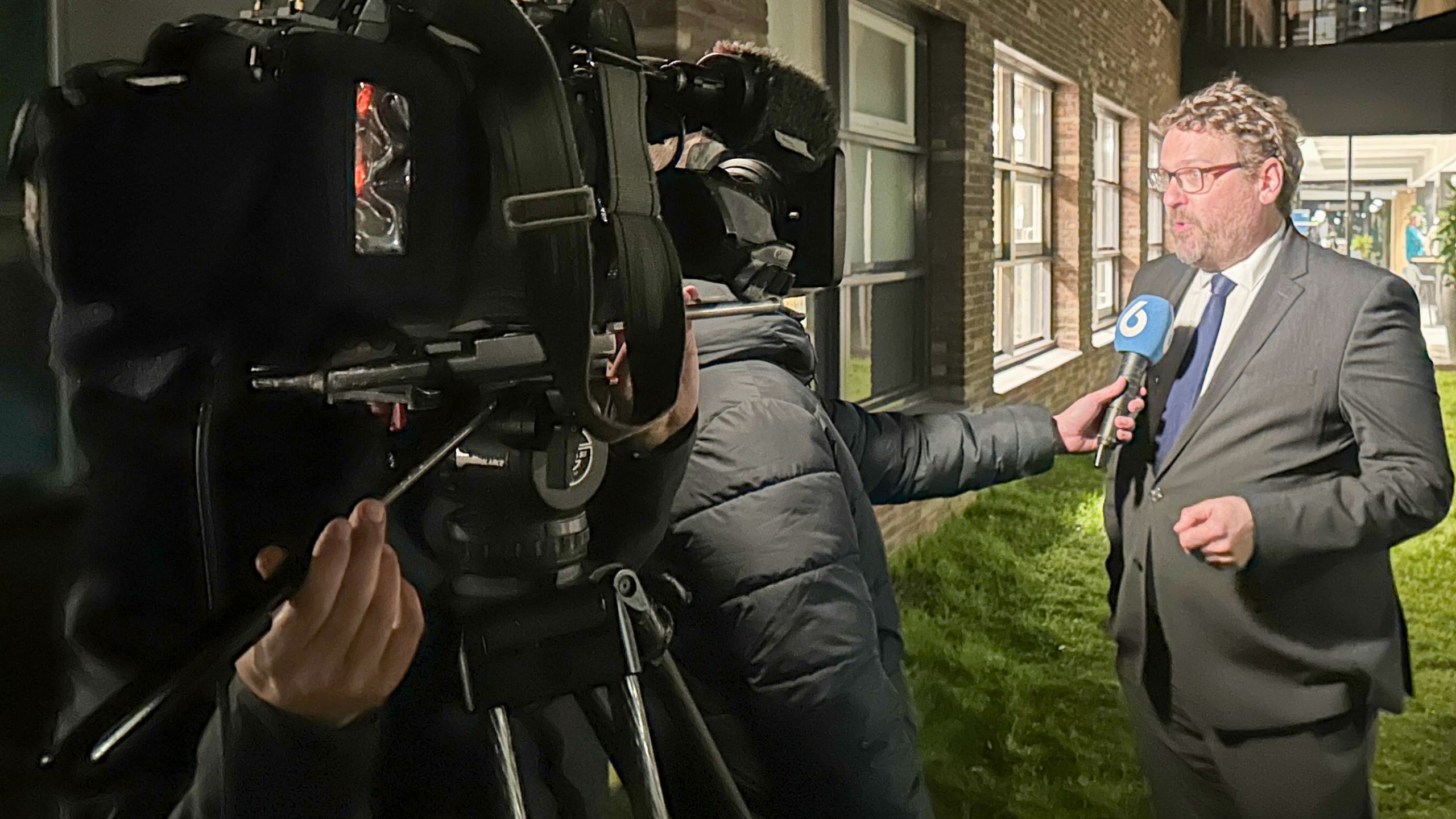Een journalist interviewt een man in pak en stropdas met een microfoon van tv-zender met nummer zes, naast een gebouw.