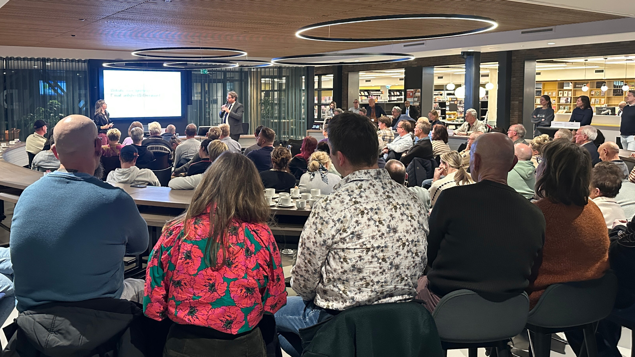 Een groep mensen zit in een moderne zaal en luistert naar sprekers die voor een scherm staan.