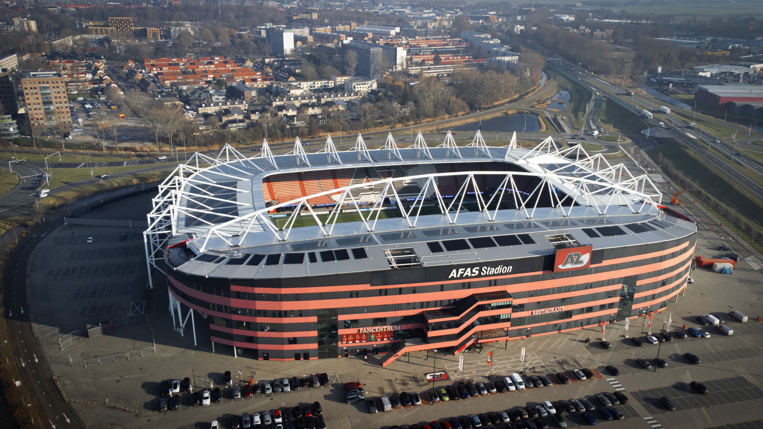 Luchtfoto van het AFAS Stadion, een groot stadion met rood-zwarte gevels en opvallende witte dakconstructie, omgeven door parkeerplaatsen.