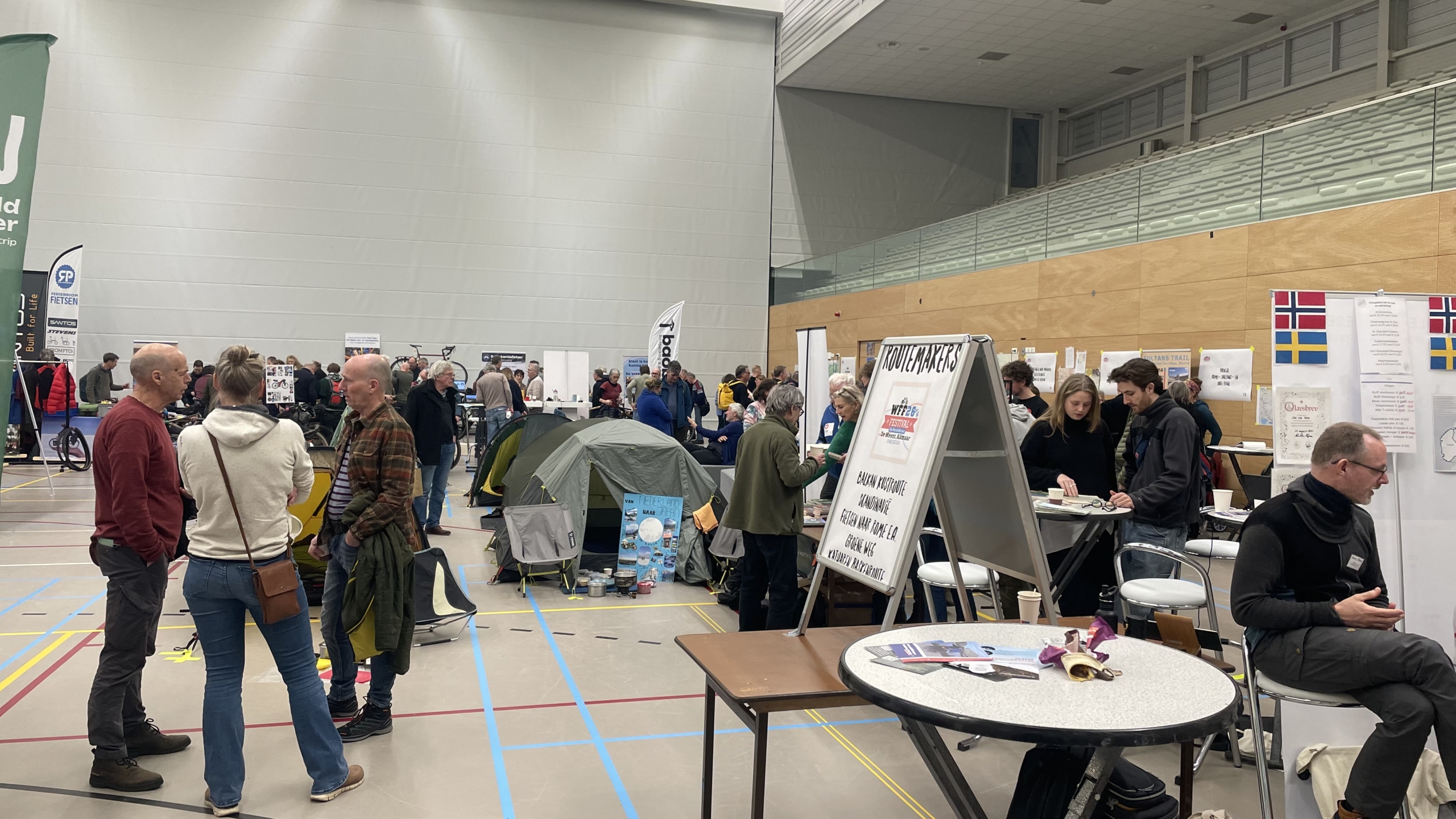 Mensen praten en navigeren door een beurs met stands en tenten, in een grote binnenruimte.