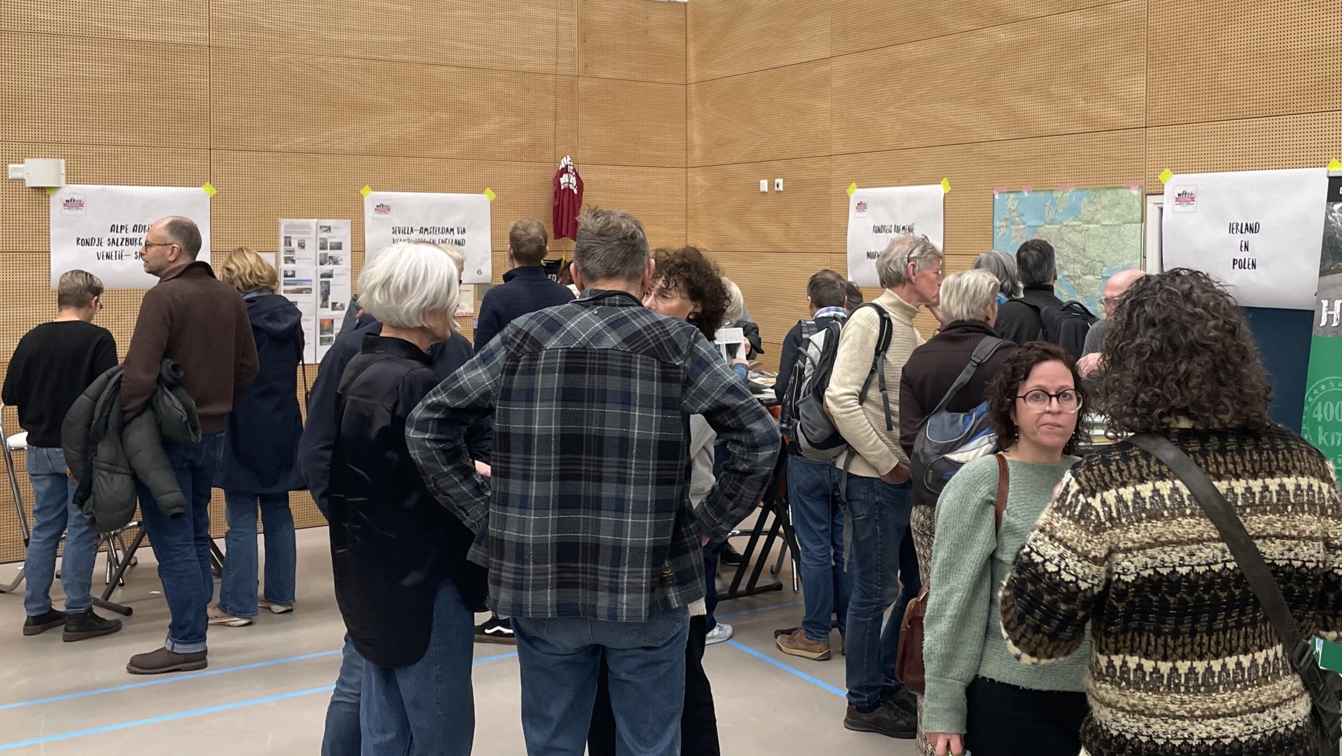 Mensen in een zaal bekijken verschillende infoposters over fietsreizen, met een poster die "De Honderd Cols Tocht" promoot.