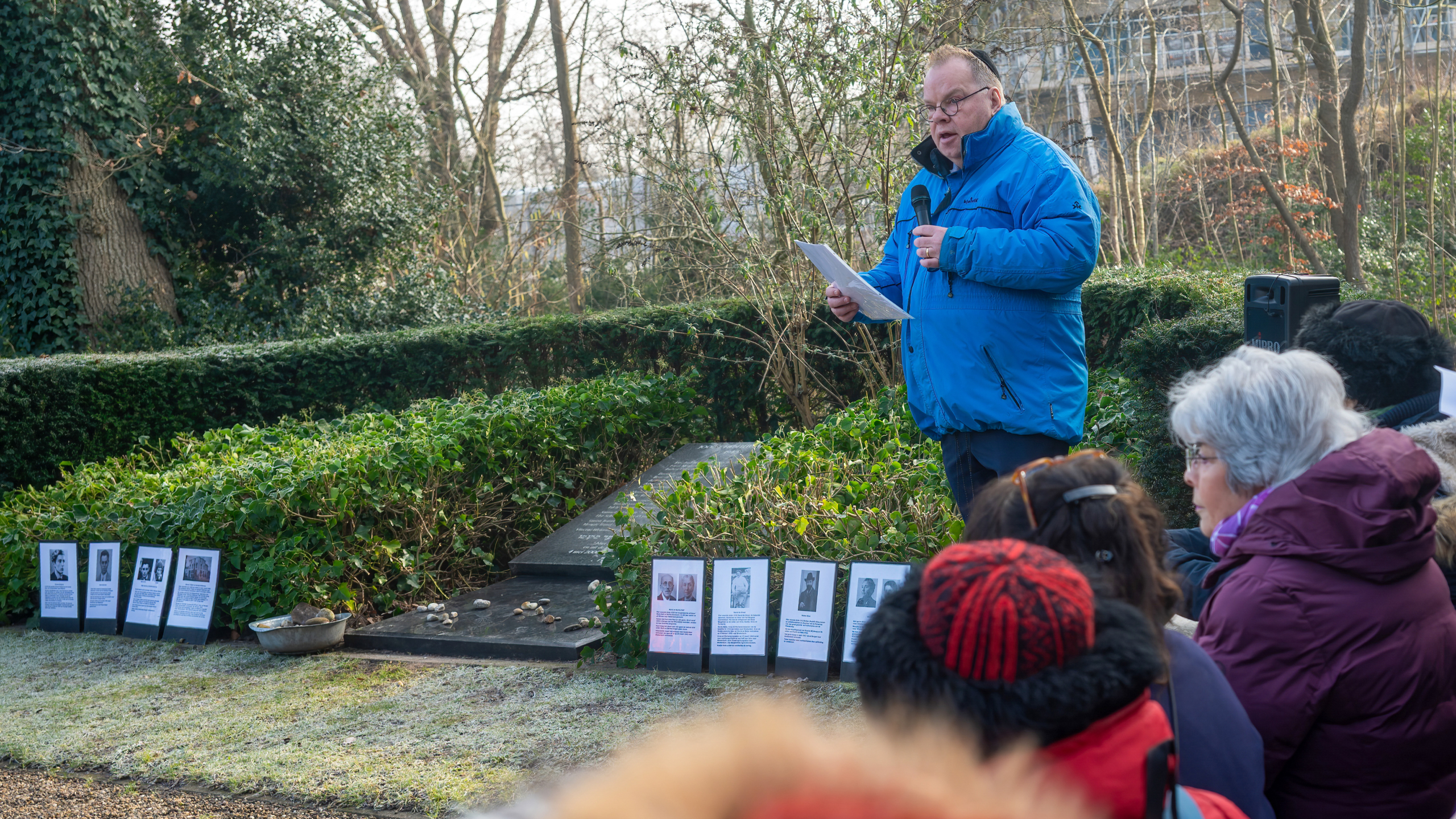 Man in een blauwe jas spreekt voor een groep mensen bij een gedenkplaats omringd door struiken en fotoborden.