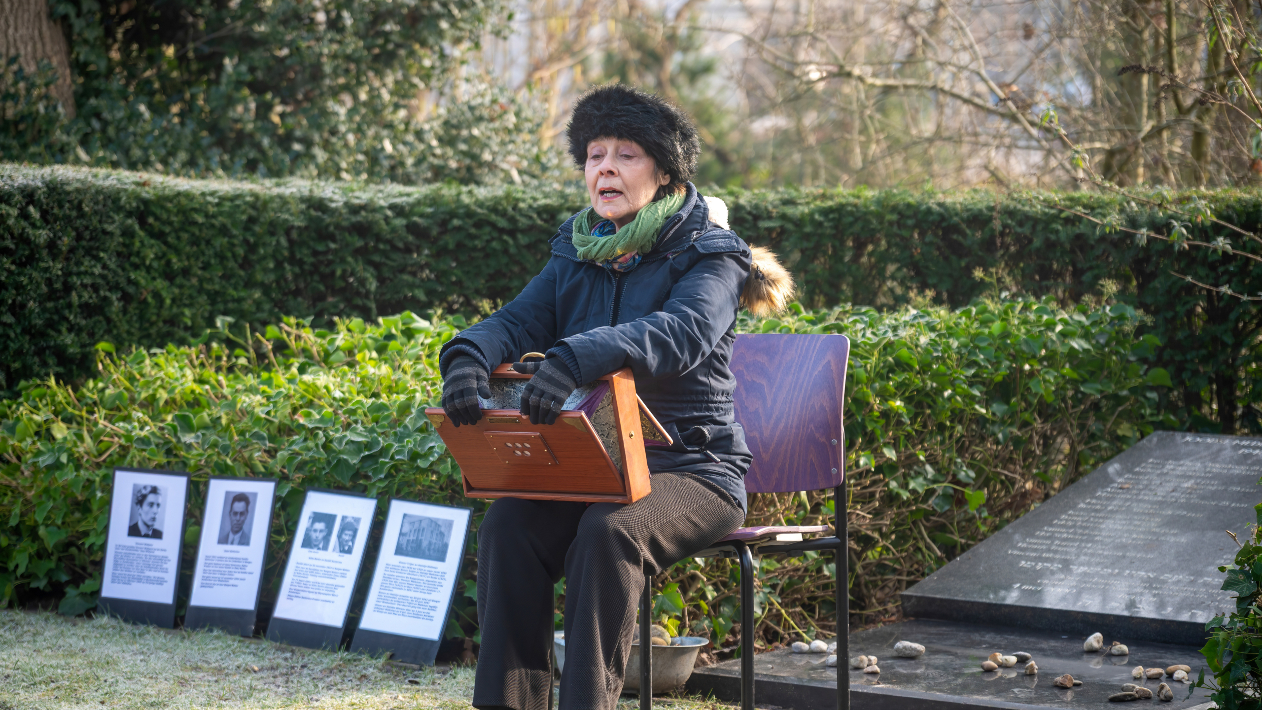 Een vrouw met winterkleding bespeelt een instrument zittend op een stoel bij een herdenkingsplek met foto's en tekst.