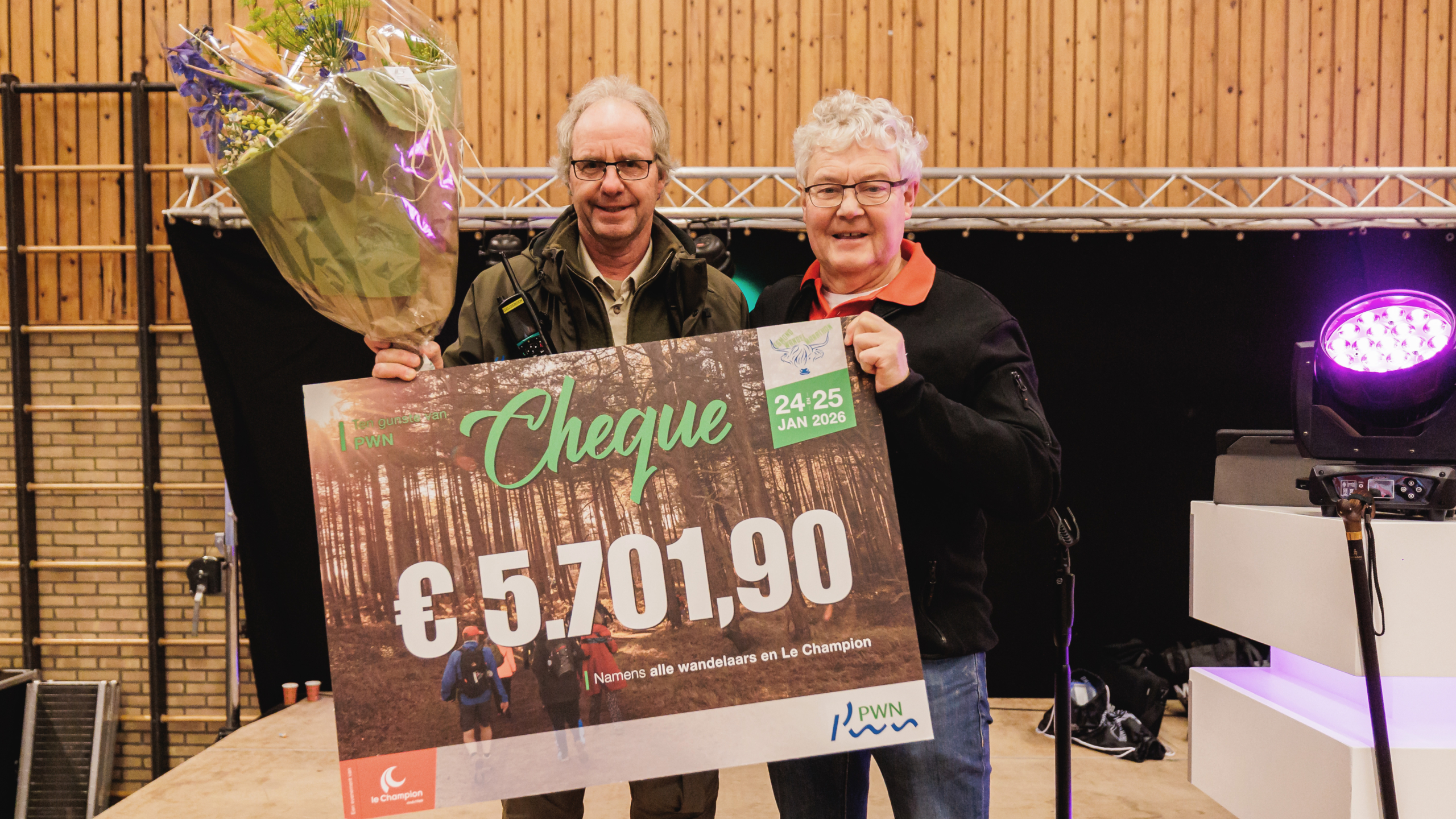 Twee mensen met een grote cheque van €5.701,90 en een boeket bloemen op een podiumsetting.