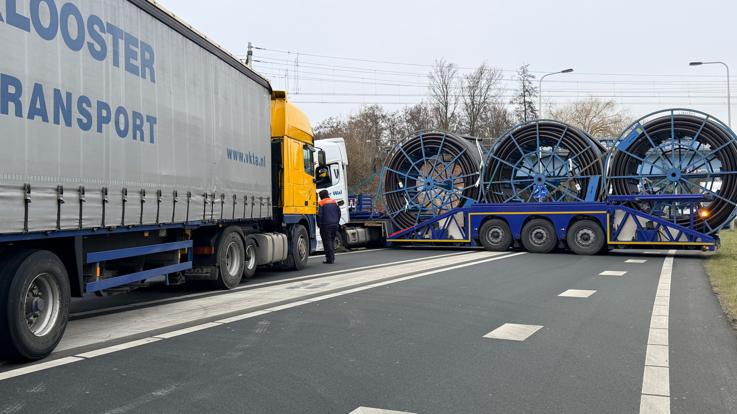 Grote vrachtwagen met spoelen op een oplegger blokkeert een weg; een persoon controleert de situatie.