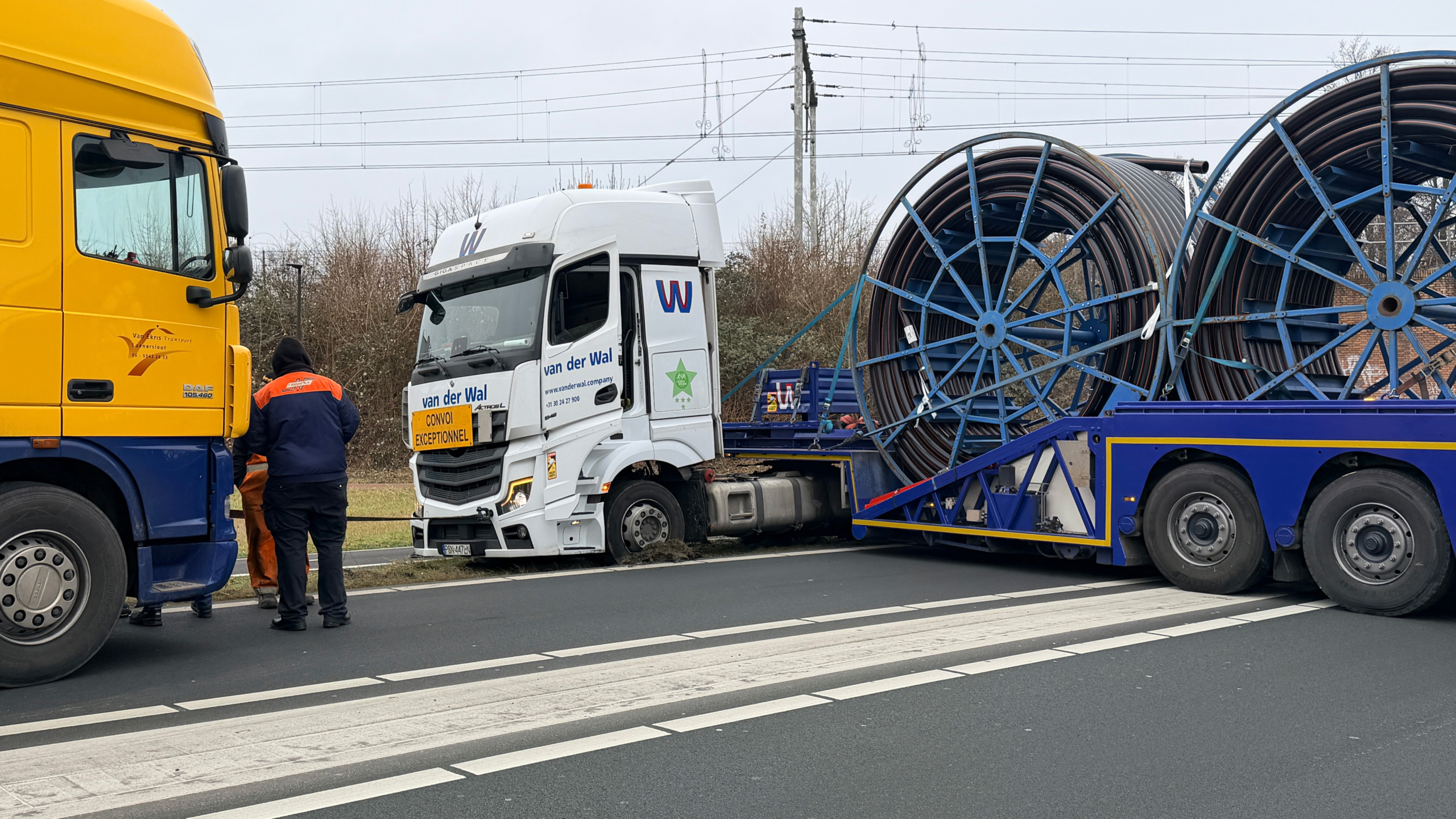Twee vrachtwagens, een gele en een witte, staan op de weg met een grote haspel op een trailer; personen ernaast.
