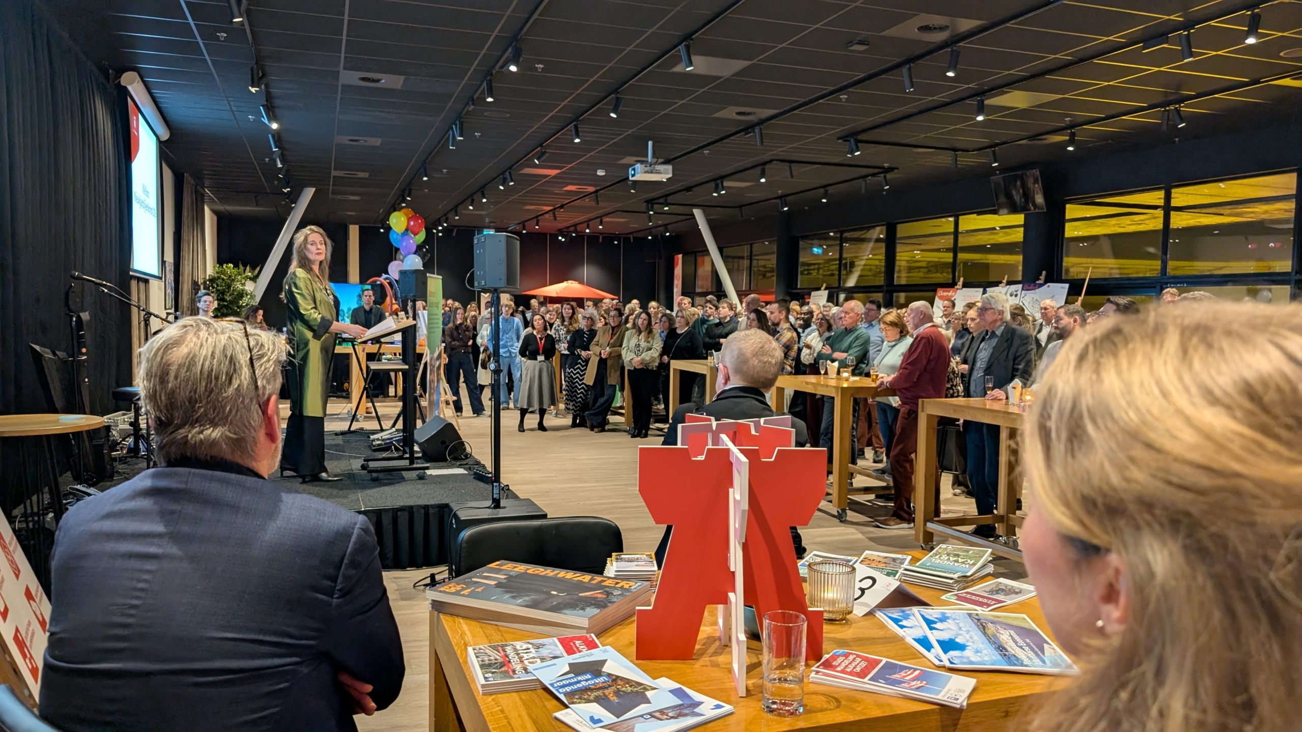 Een vrouw staat op een podium en spreekt voor een zaal vol mensen tijdens een evenement.