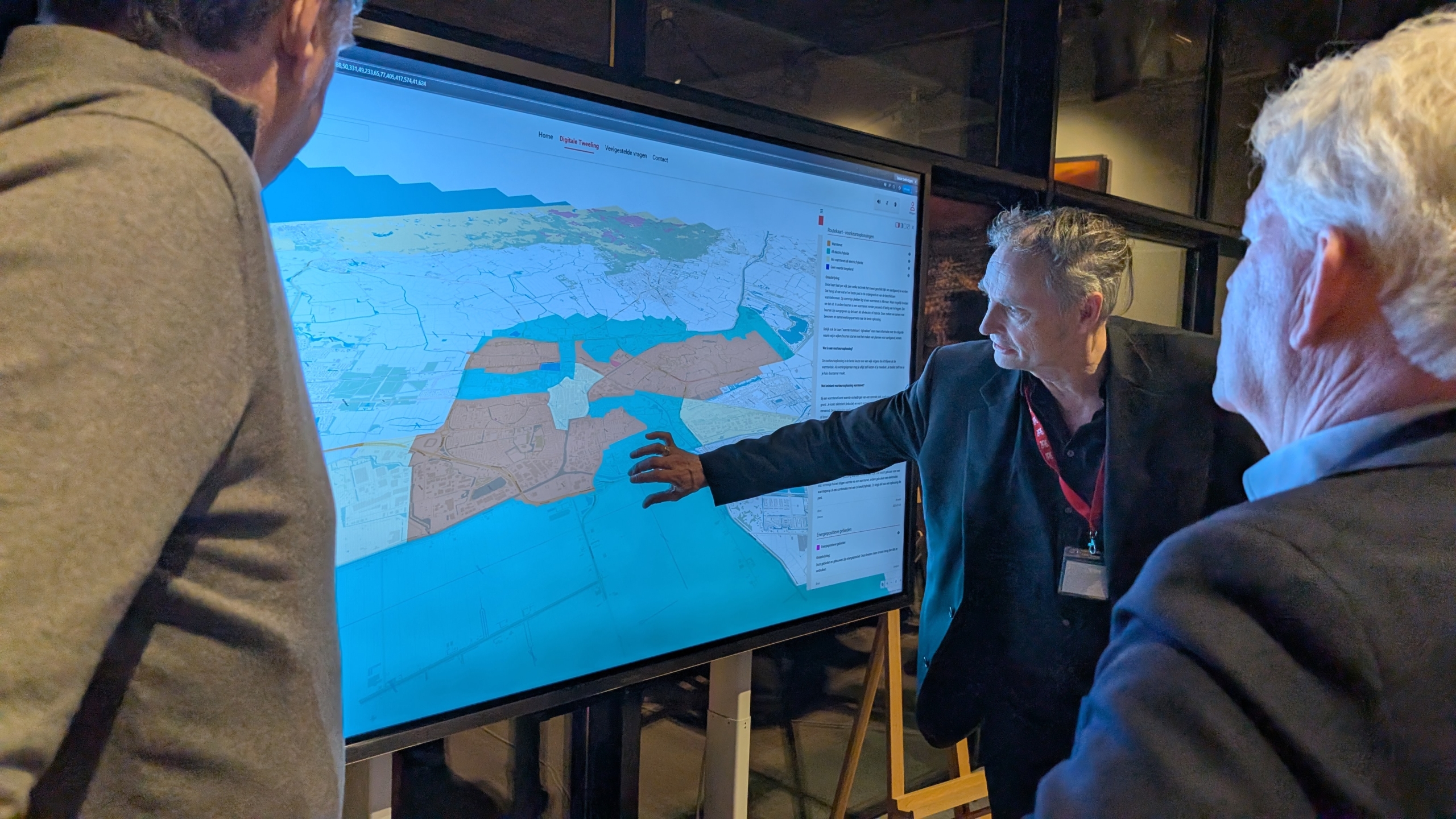 Drie mannen bekijken een kaart op een groot scherm, terwijl een van hen iets aanwijst op de kaart.