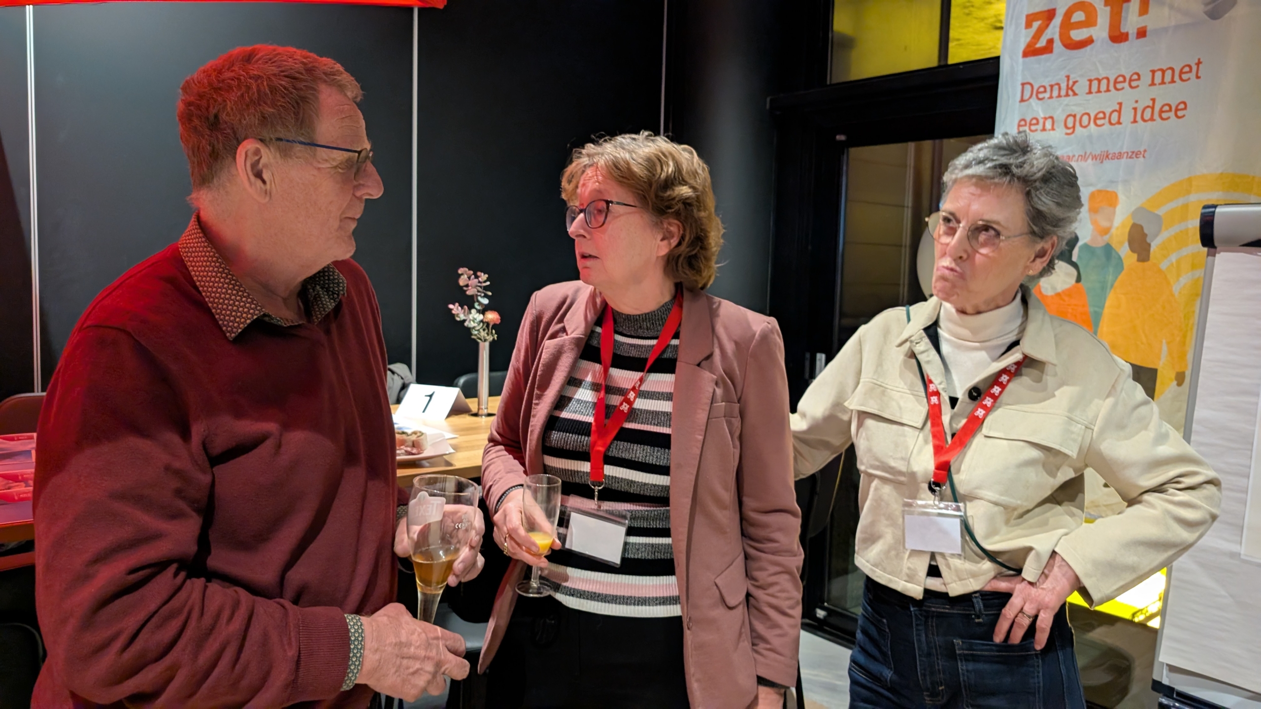 Drie mensen in gesprek bij een tafel met glazen; op de achtergrond een bord met de tekst "Denk mee met een goed idee".