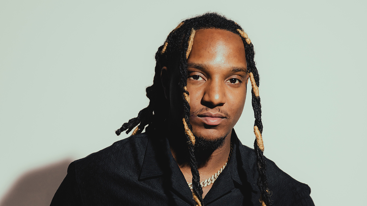 Man met lang haar deels blond, draagt een zwart overhemd en zilveren ketting, tegen een lichte achtergrond.