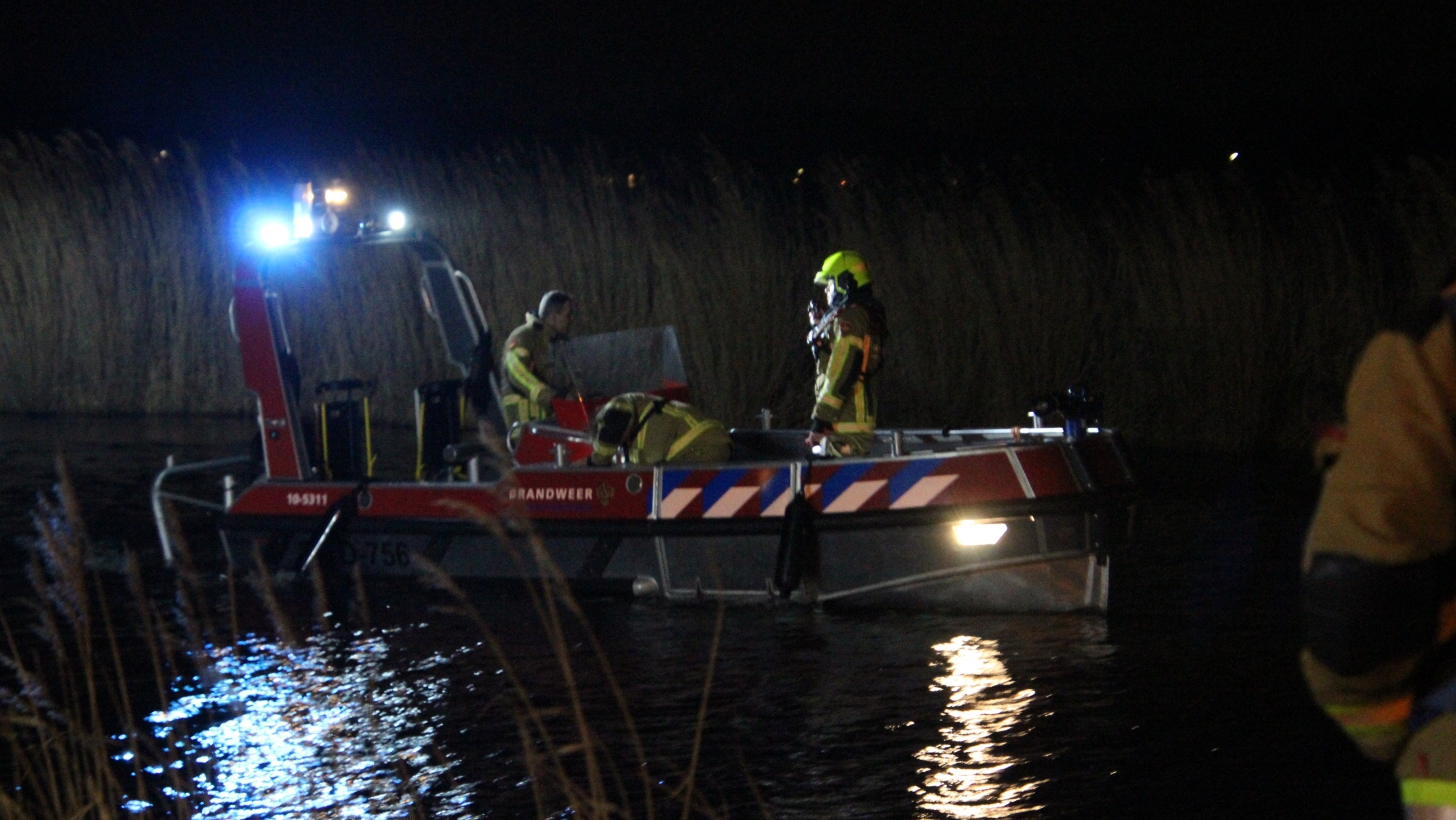 Brandweerlieden op een reddingsboot uitvoeren een nachtelijke operatie op een meer, verlicht door felle lichten.