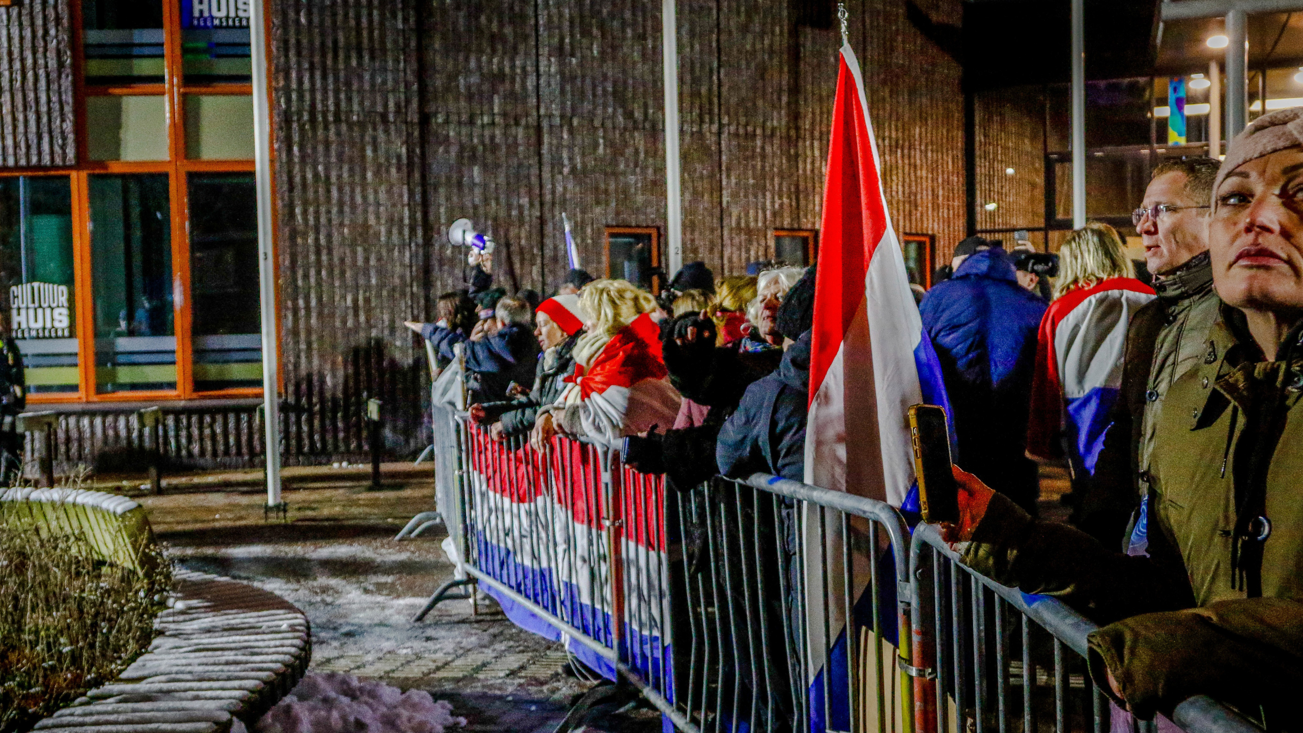 Mensen staan achter dranghekken met een Nederlandse vlag en spandoek, bij een gebouw met de tekst "Cultuur Huis".