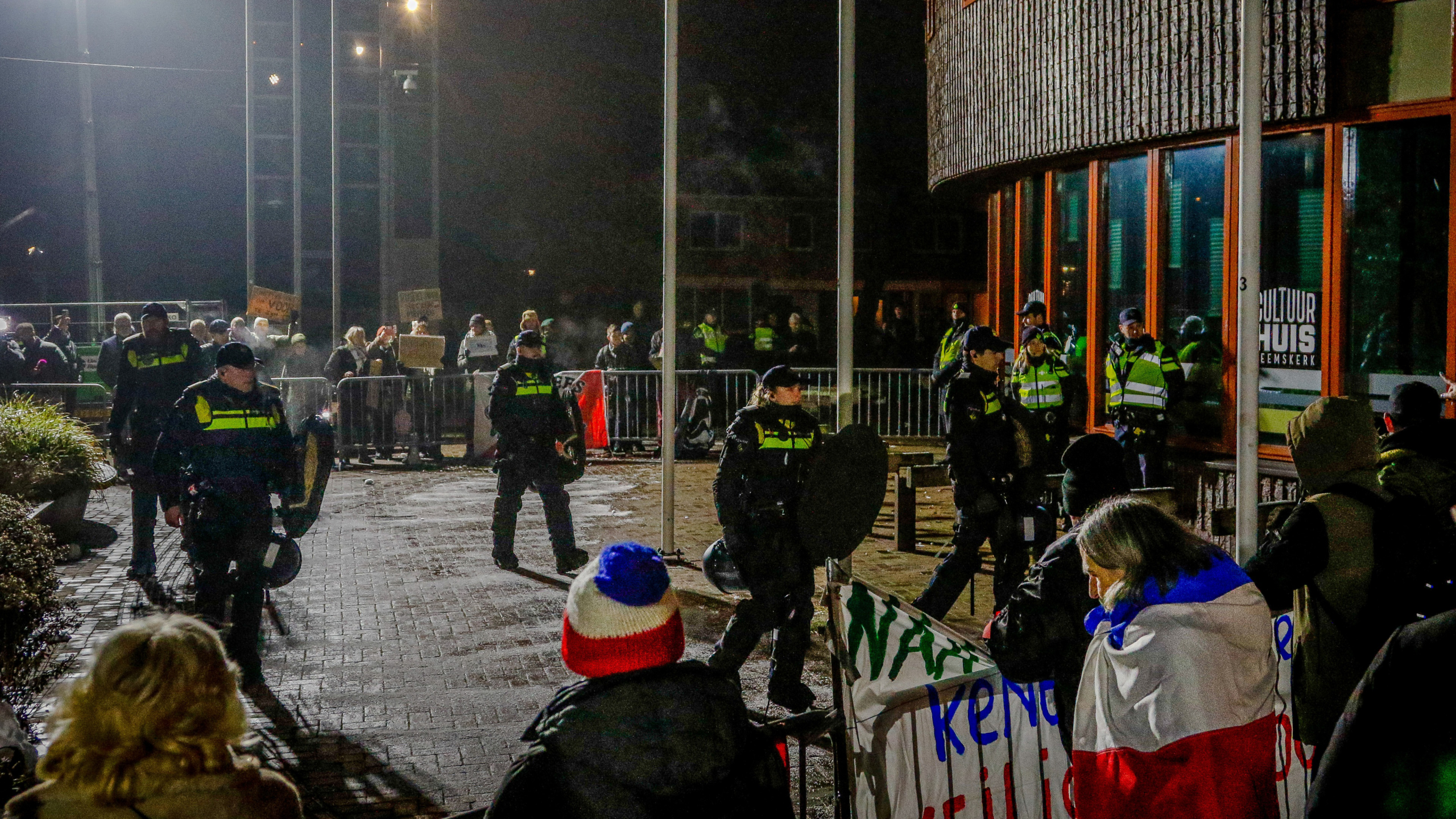 Politieagenten staan voor een gebouw; een menigte met borden bevindt zich achter dranghekken.