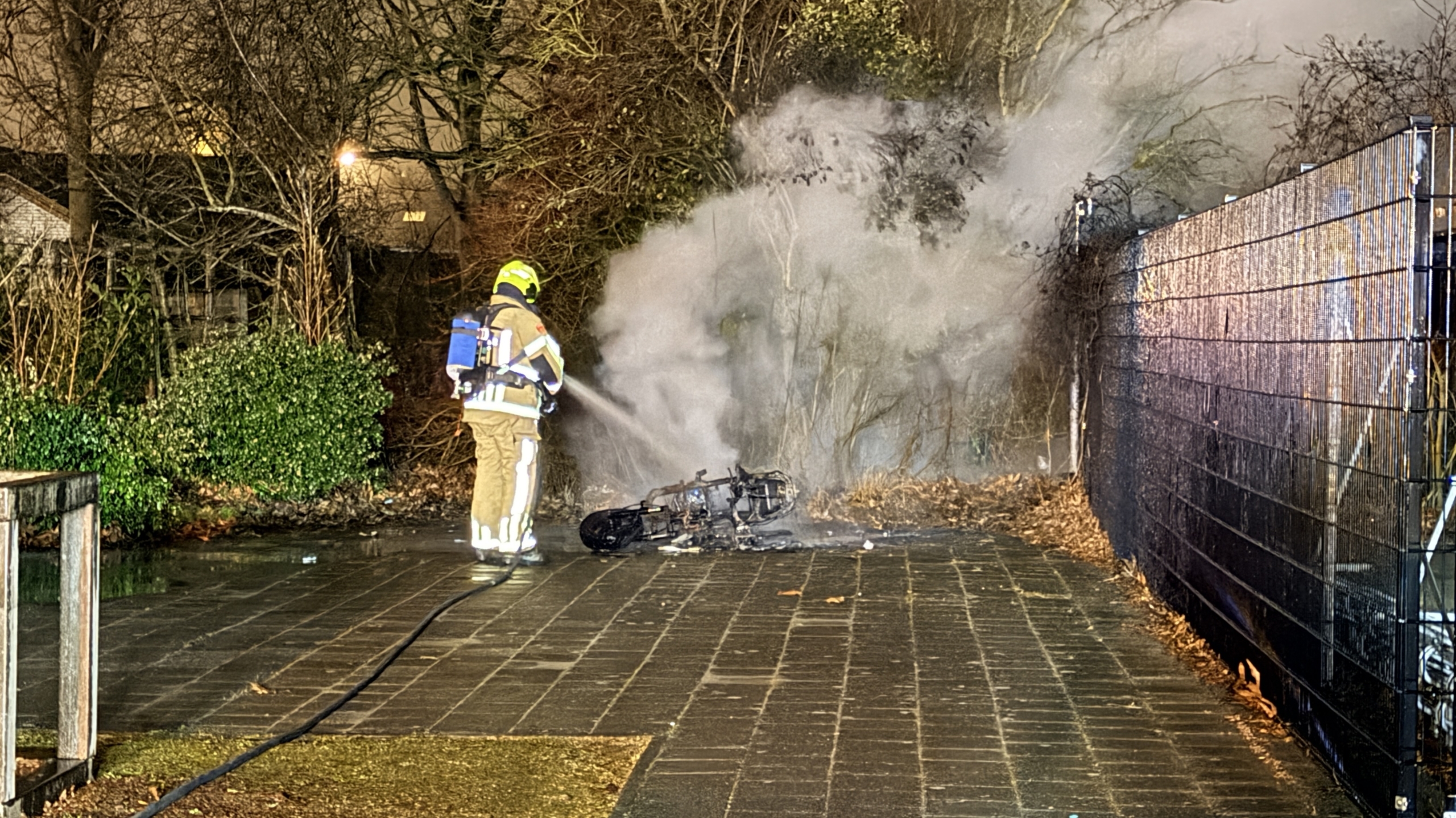 Brandweerman blust brandende scooter langs een pad, omgeven door rook en bomen.