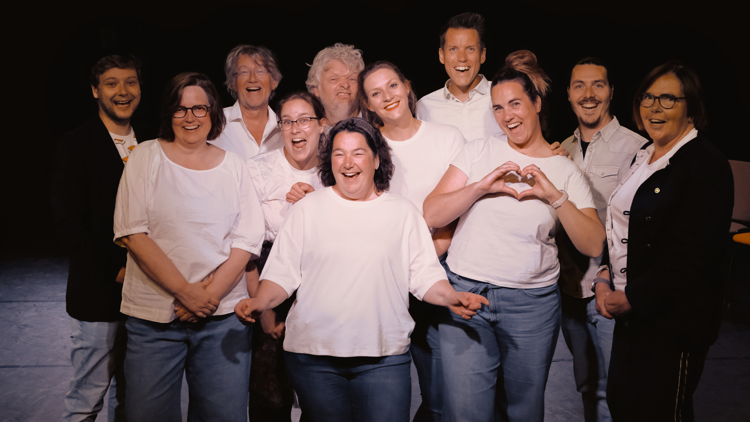 Een groep van elf personen poseert lachend voor de camera, sommigen dragen witte bovenkleding, een persoon maakt een hartvorm met de handen.