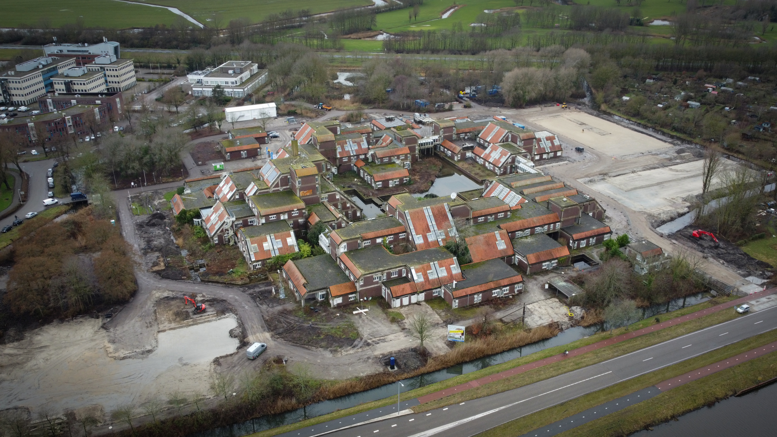 Luchtfoto van een woonwijk met rode daken, omgeven door wegen, water en groen, met een bouwplaats en graafmachine.