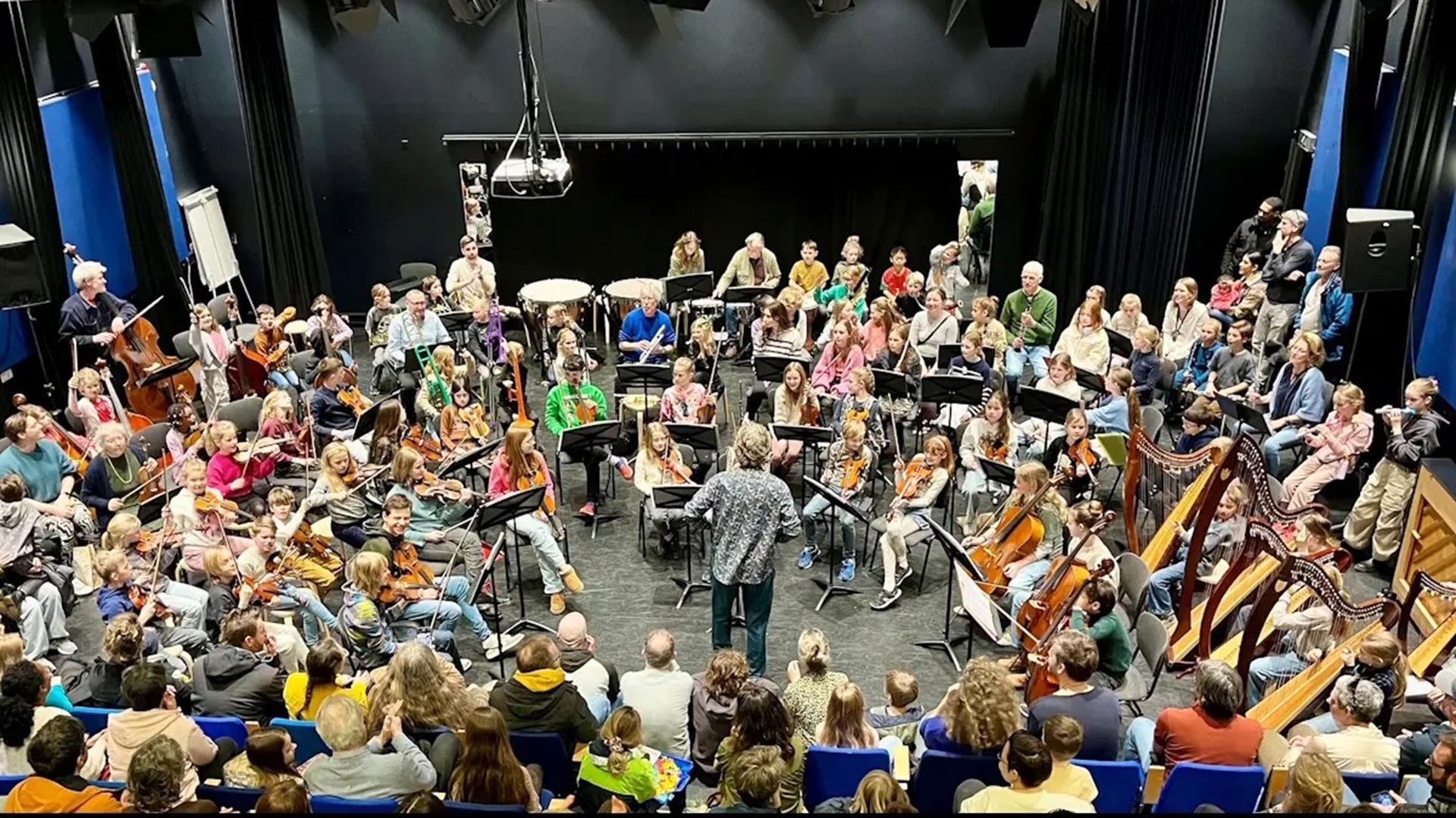 Een orkest van kinderen en volwassenen speelt muziek samen in een concertzaal, onder leiding van een dirigent.