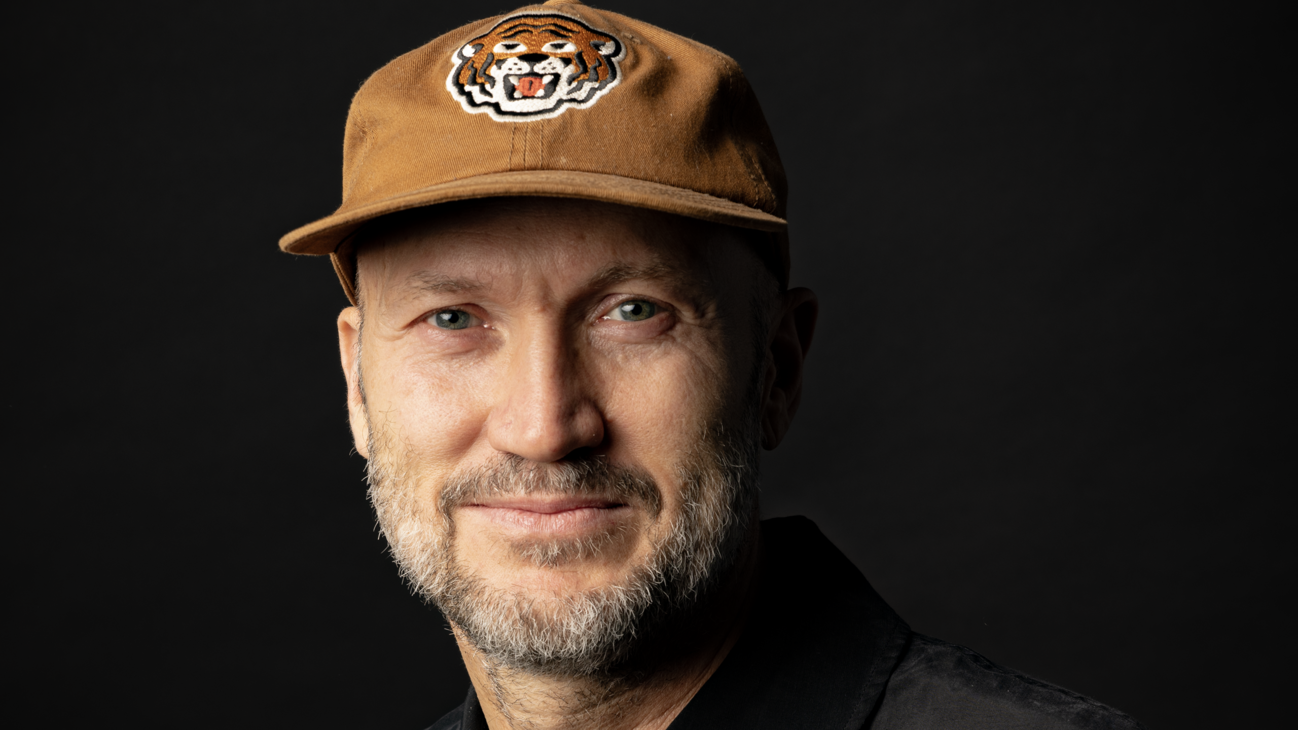Man met baard en snor draagt een bruine pet met een tijgerlogo, tegen een donkere achtergrond.