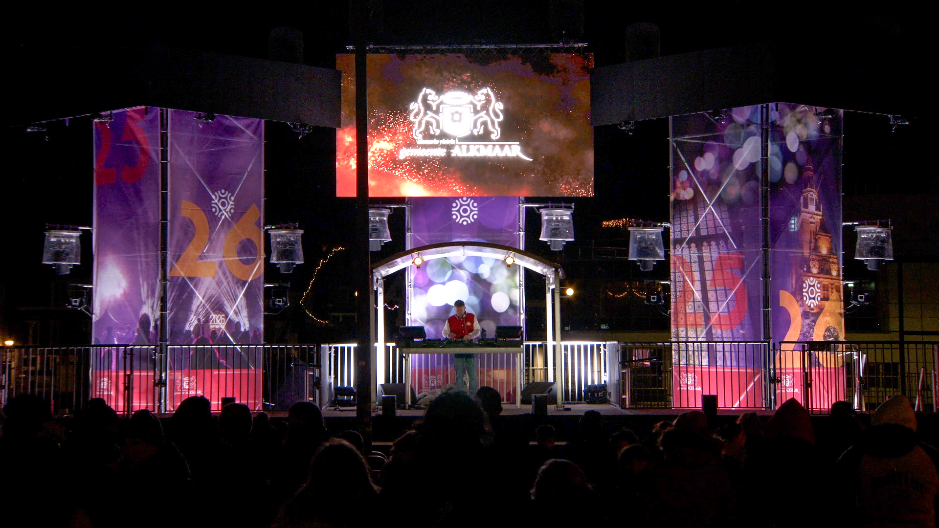 Podium in Alkmaar met een dj die muziek draait voor een publiek bij nacht, versierd met kleurrijke banners en een groot scherm.