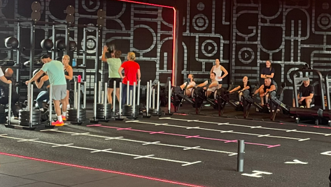 Mensen trainen in een sportschool, bezig met verschillende fitnessapparaten, tegen een achtergrond met een opvallend patroon.