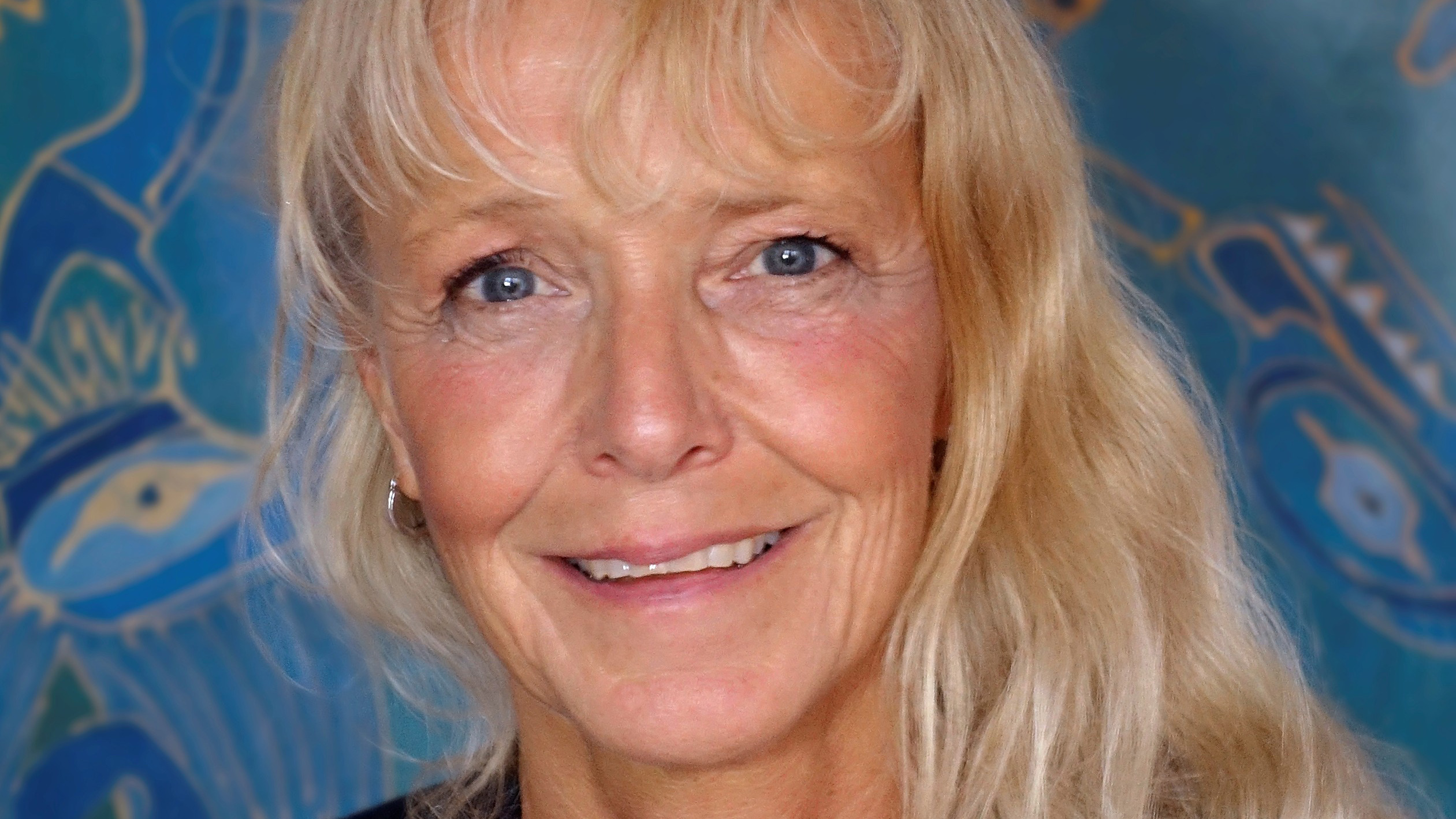 Blonde vrouw met een vriendelijke glimlach, voor een blauwe achtergrond met abstracte patronen.