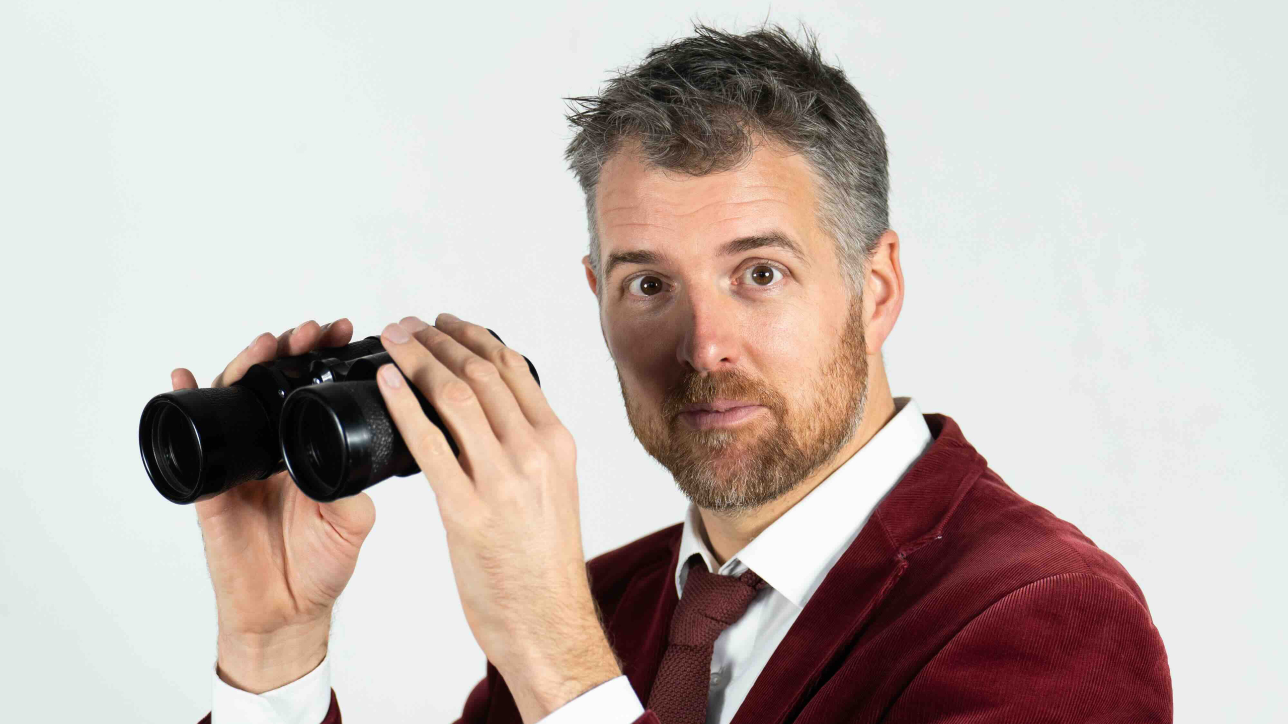 Man met een baard en een rood fluwelen jasje houdt een verrekijker vast en kijkt recht vooruit.