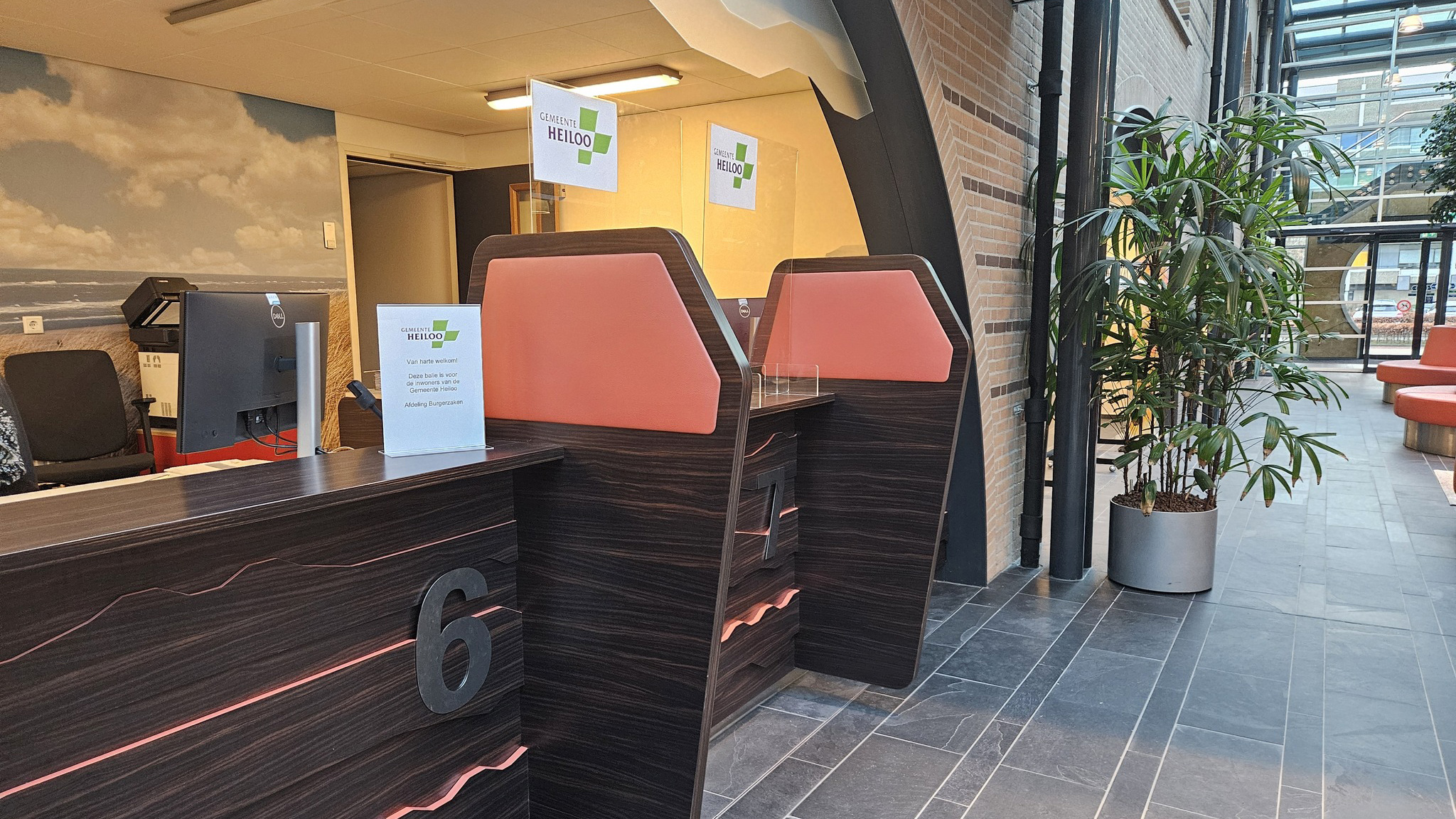 Interieur van een gemeentekantoor met een ontvangstbalie, stoelen, een computer en plant.