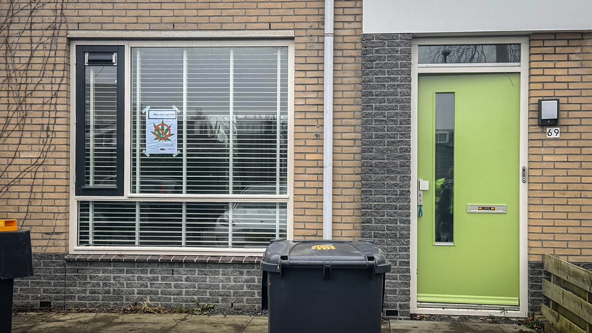 Een huis met een felgroene voordeur en een groot raam met jaloezieën, met een poster op het raam. Een zwarte vuilnisbak staat op de stoep.
