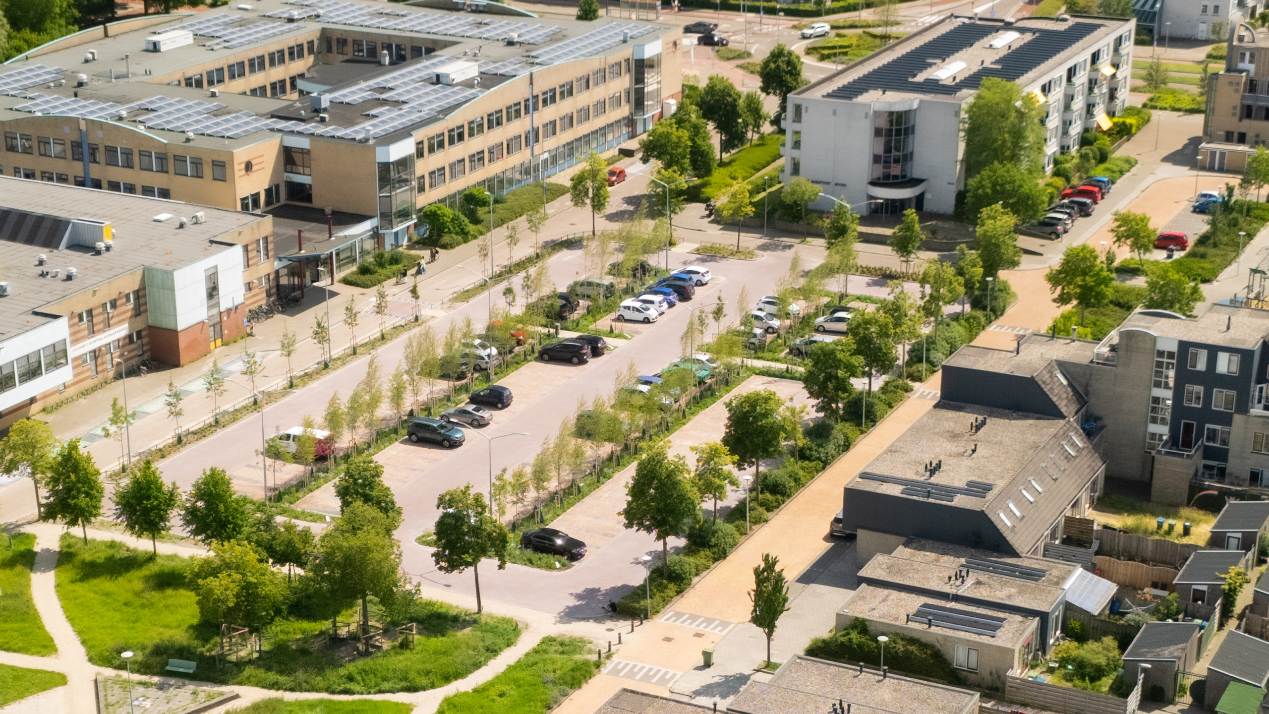 Parkeerplaats met auto's naast een groot gebouw met zonnepanelen op het dak, omringd door groen en andere gebouwen.
