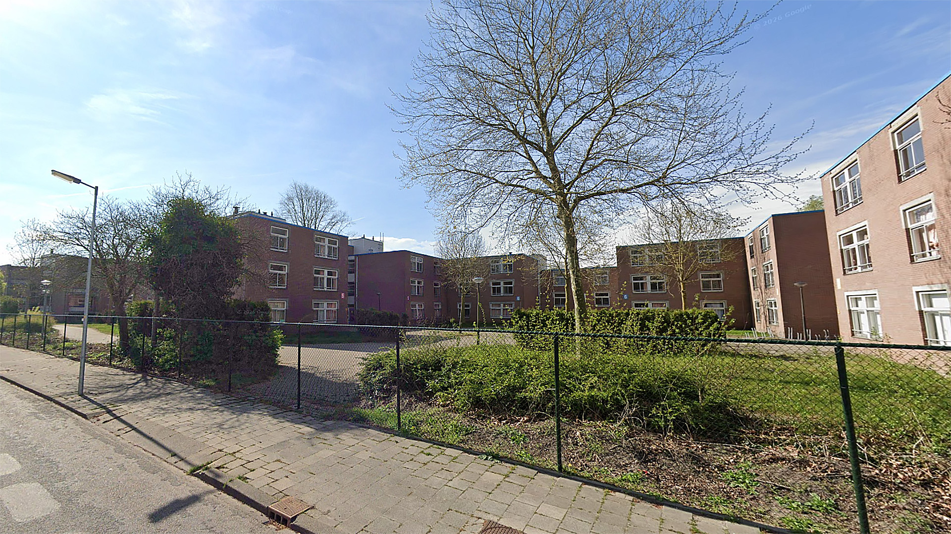 Appartementencomplex met drie verdiepingen en rode bakstenen, omgeven door een hek en bomen, onder een blauwe lucht.