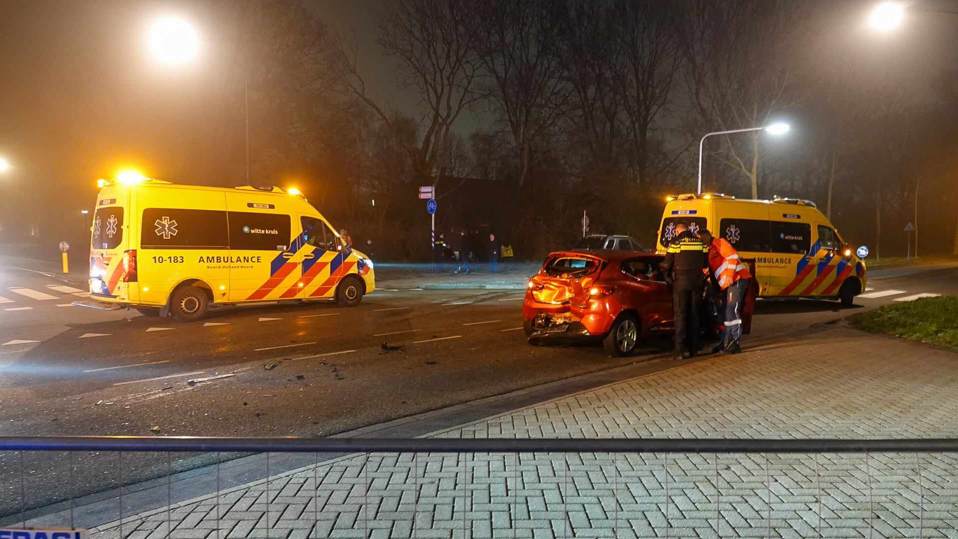 Twee ambulances en hulpverleners staan bij een rode auto met schade na een ongeval op een kruispunt 's nachts.