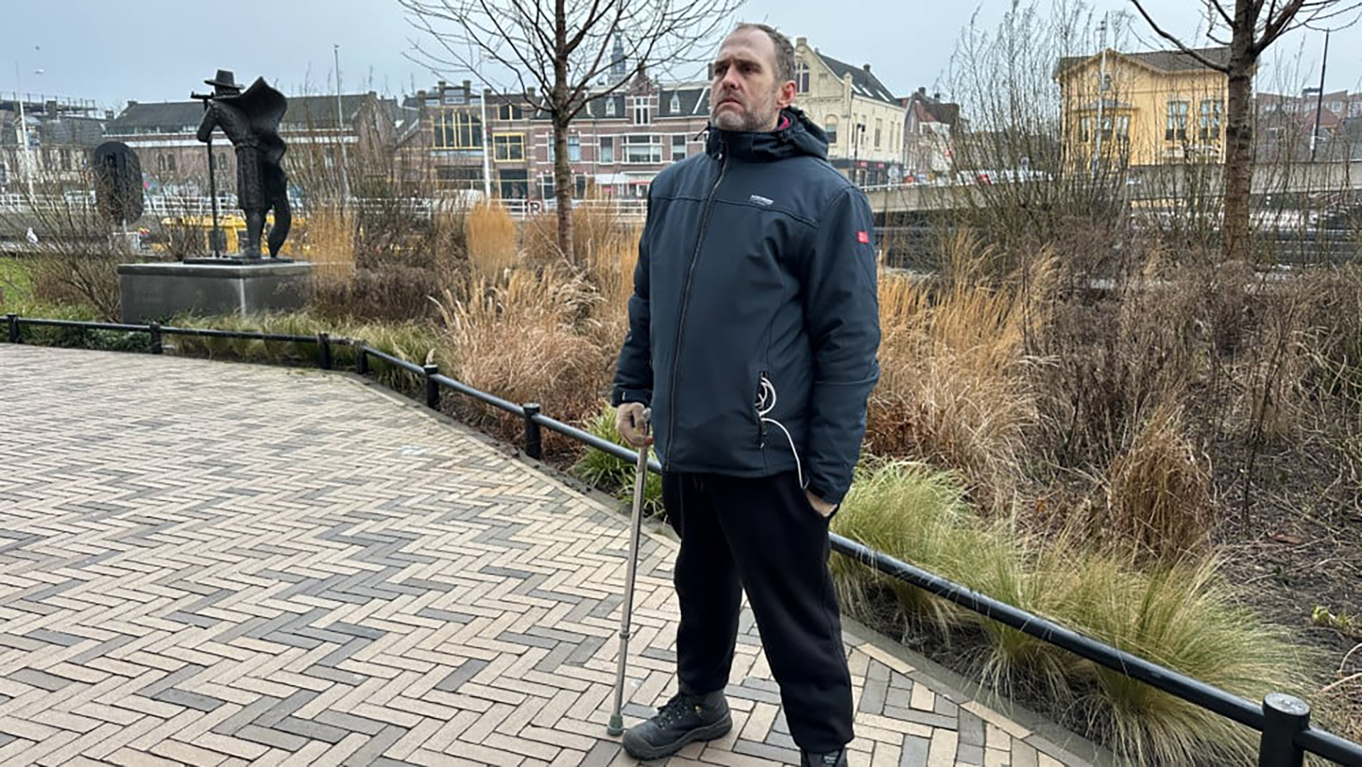 Man met een donkere jas en wandelstok staat buiten op een geplaveid pad, met struiken en een standbeeld op de achtergrond.