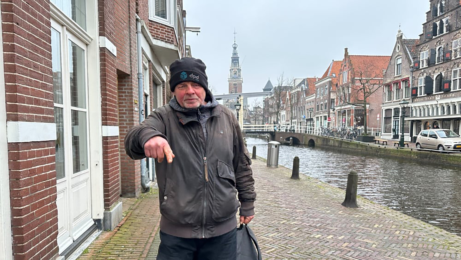 Man in winterkleding staat naast een gracht met historische gebouwen op de achtergrond.