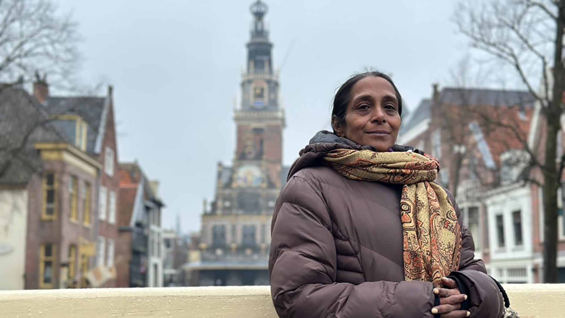 Vrouw in warme jas met sjaal staat voor historische gebouwen en een toren tegen een grijze lucht.