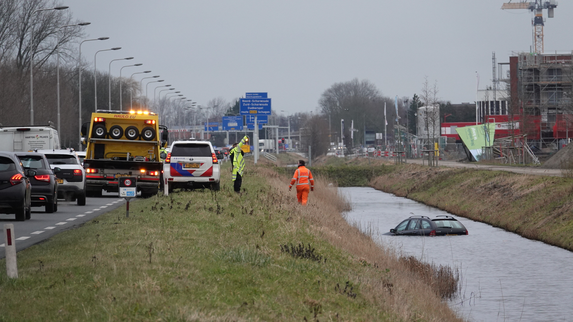 Auto deels ondergedompeld in een sloot langs een weg, met hulpverleners en politievoertuig aanwezig.