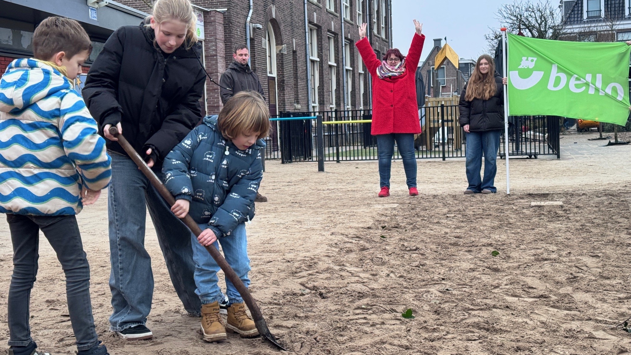 Kinderen graven in een zanderige speelplaats terwijl een vrouw juicht en een meisje een groene vlag vasthoudt.