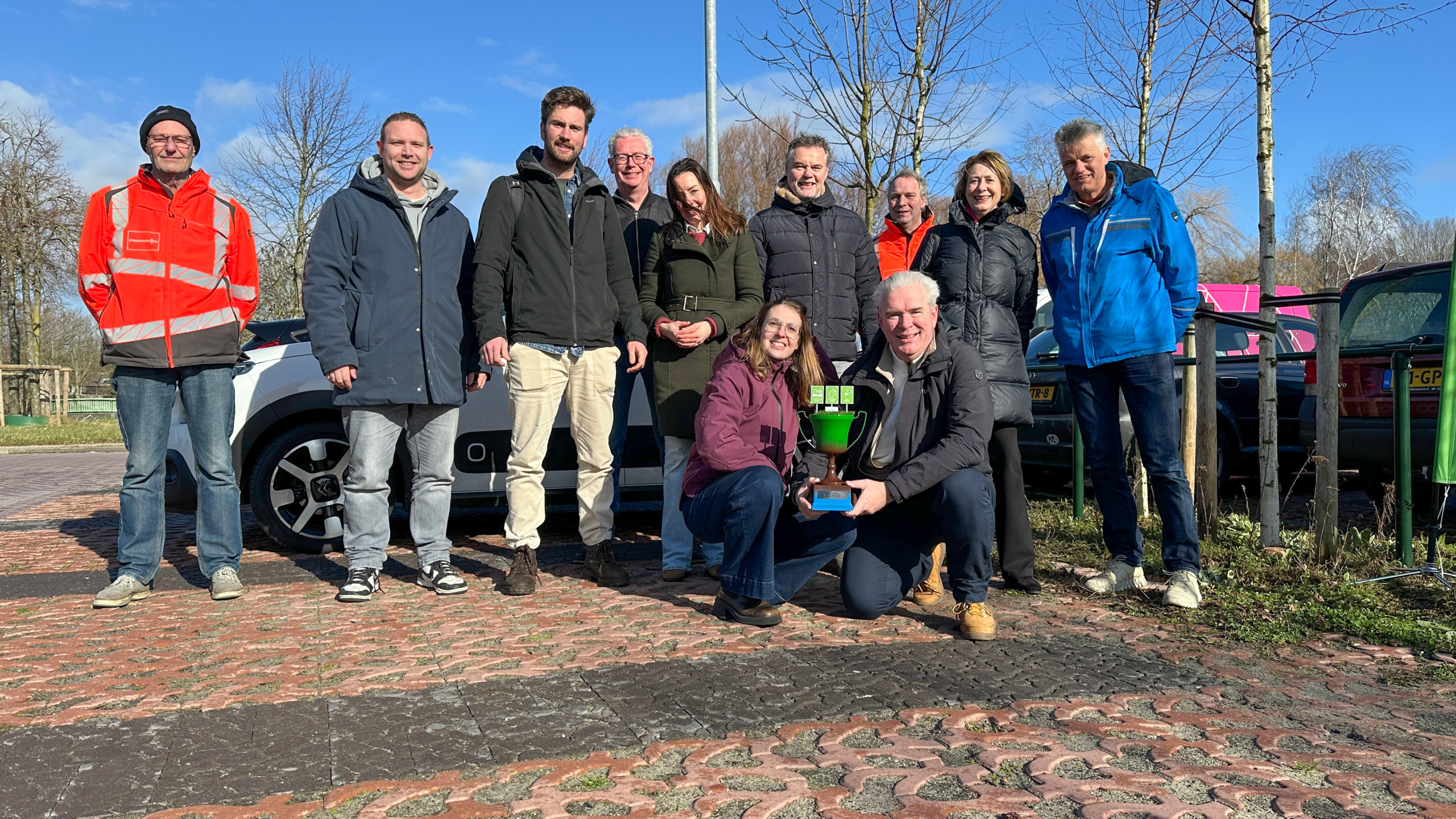Groep mensen poseren buiten met een groene trofee op een zonnige dag.