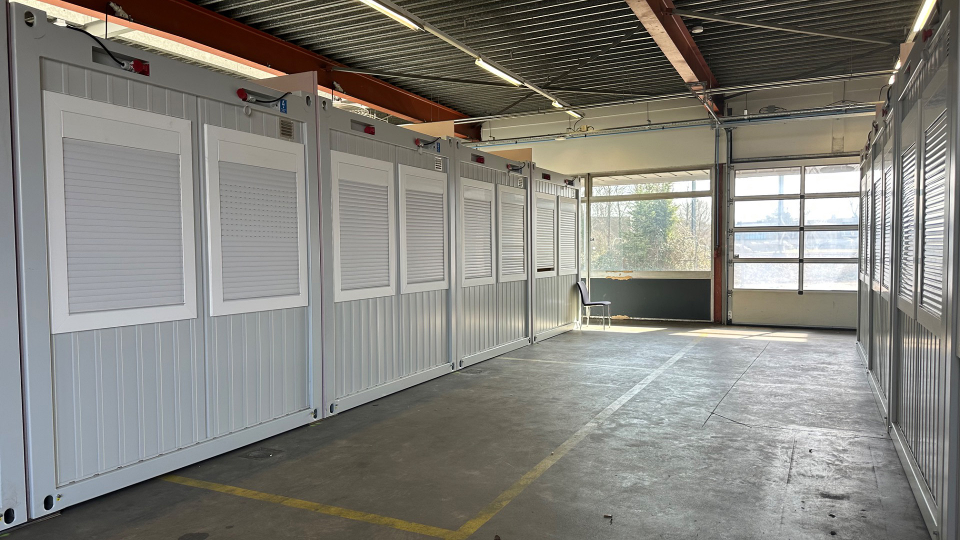 Rij van grijze containerunits in een grote, lege loods met een glazen wand en een enkele stoel.