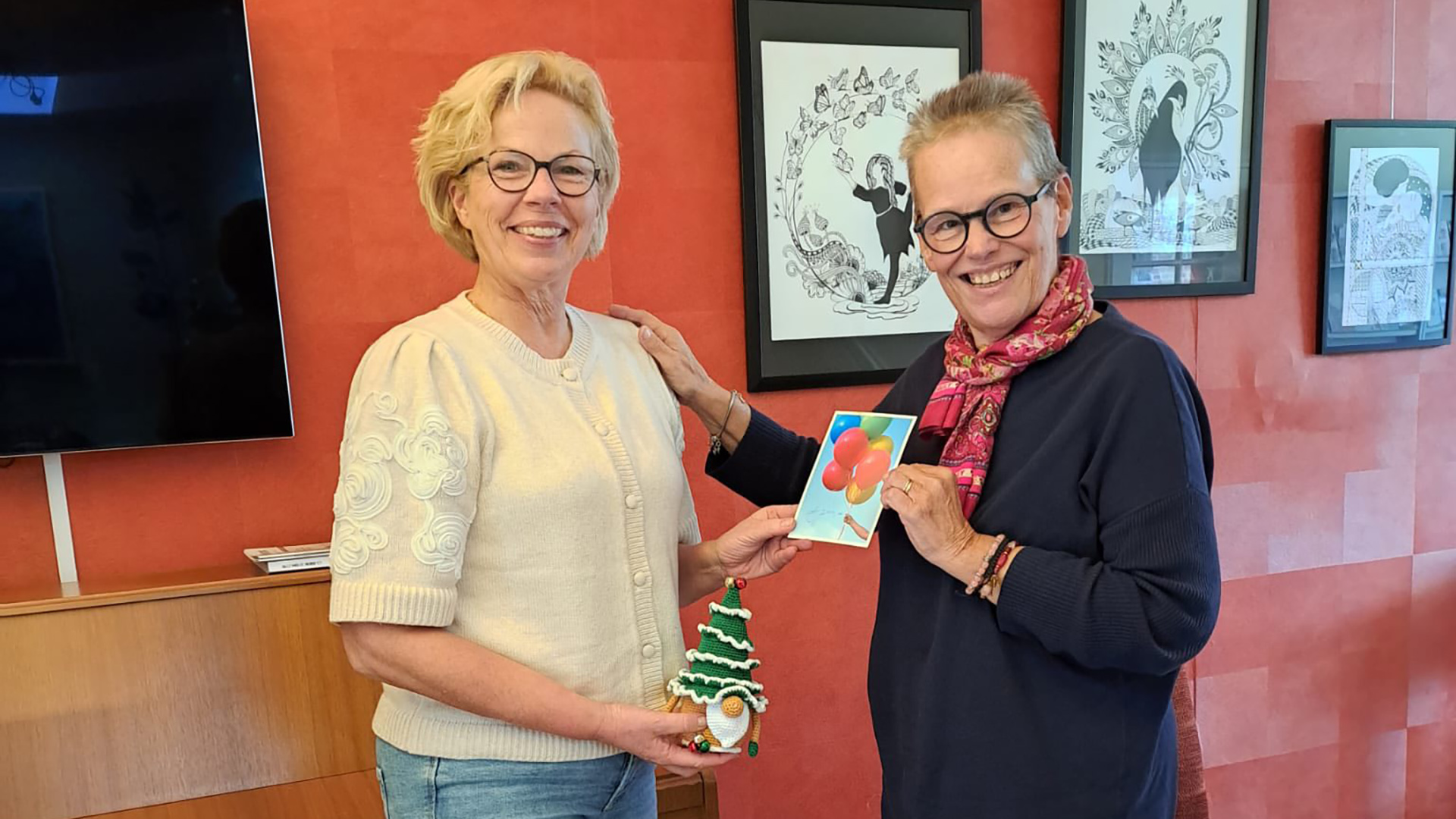 Twee vrouwen staan binnen met een kaart en een gehaakte kerstboomknuffel, glimlachend naar de camera.