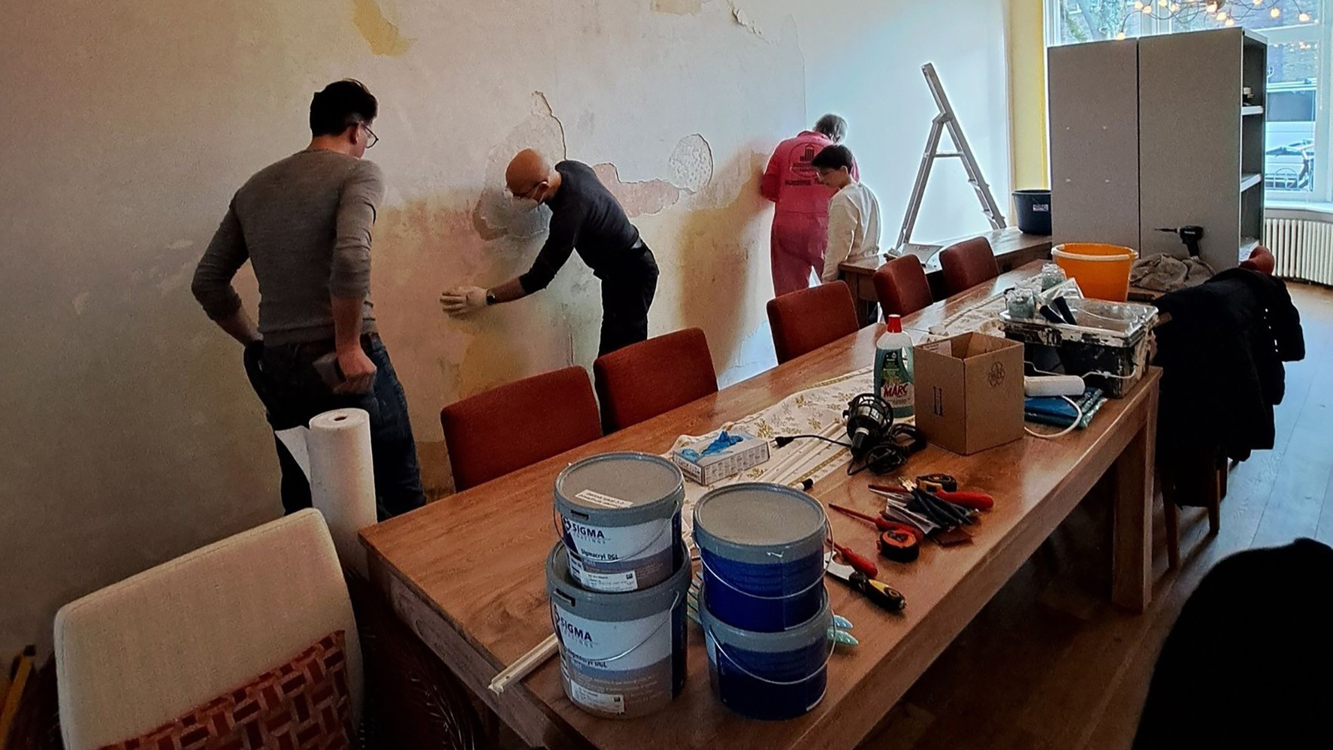 Mensen werken aan een muur in een ruimte met stoelen, tafels vol gereedschap en potten verf.