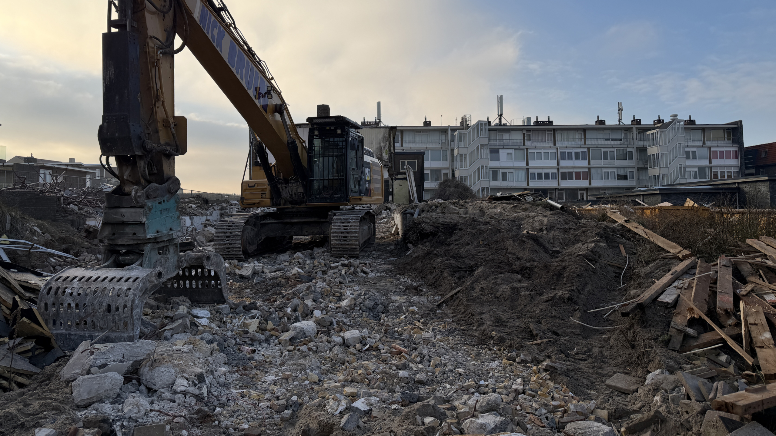 Graafmachine op een bouwplaats te midden van puin, met op de achtergrond een appartementengebouw onder een bewolkte hemel.