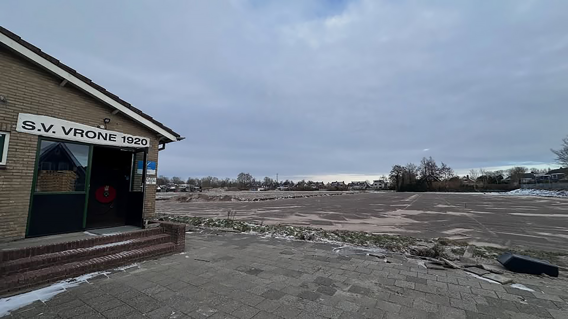 Voetbalclubgebouw met opschrift "S.V. Vrone 1920" naast een besneeuwd, leeg sportveld bij bewolkte hemel.