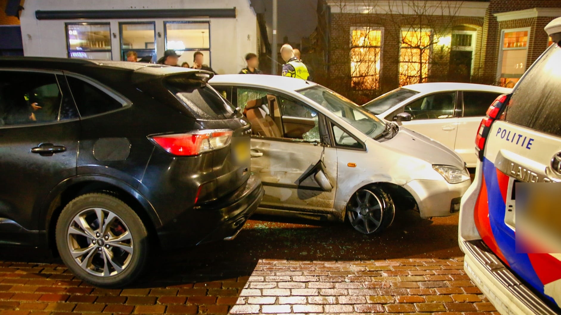Drie auto's zijn betrokken bij een botsing op een natte straat, met een politieauto en agenten op de achtergrond.